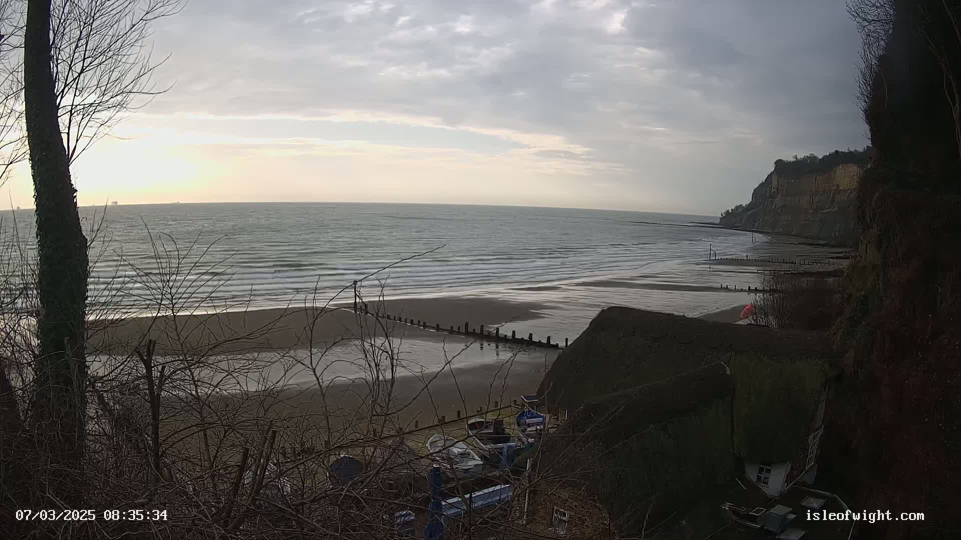 A sandy beach with gentle waves under a cloudy sky is seen from a high vantage point, with several small boats pulled ashore near a cliffside structure.