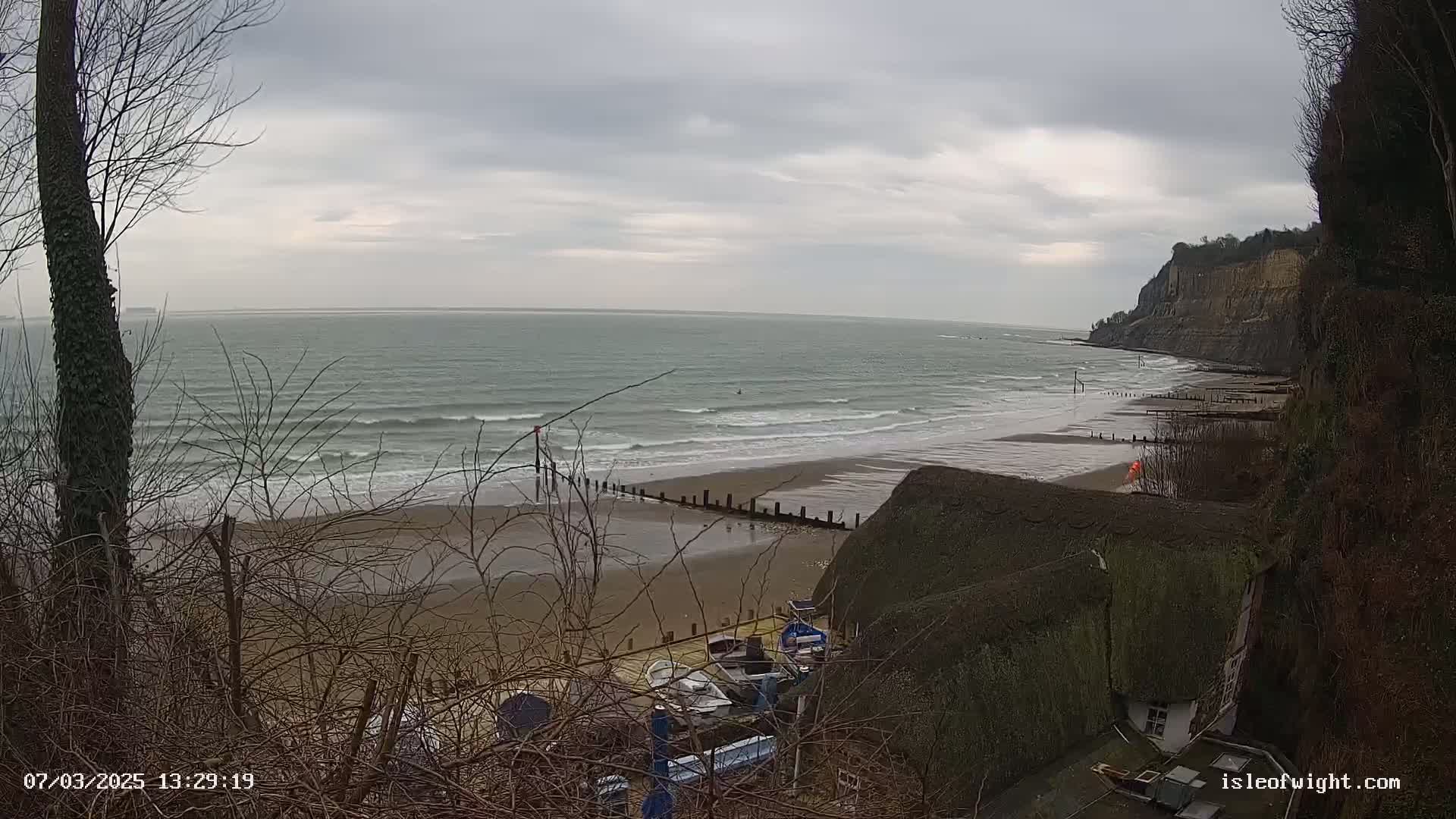 A view from a clifftop shows a beach with small waves under an overcast sky, boats pulled ashore near a thatched-roof building.