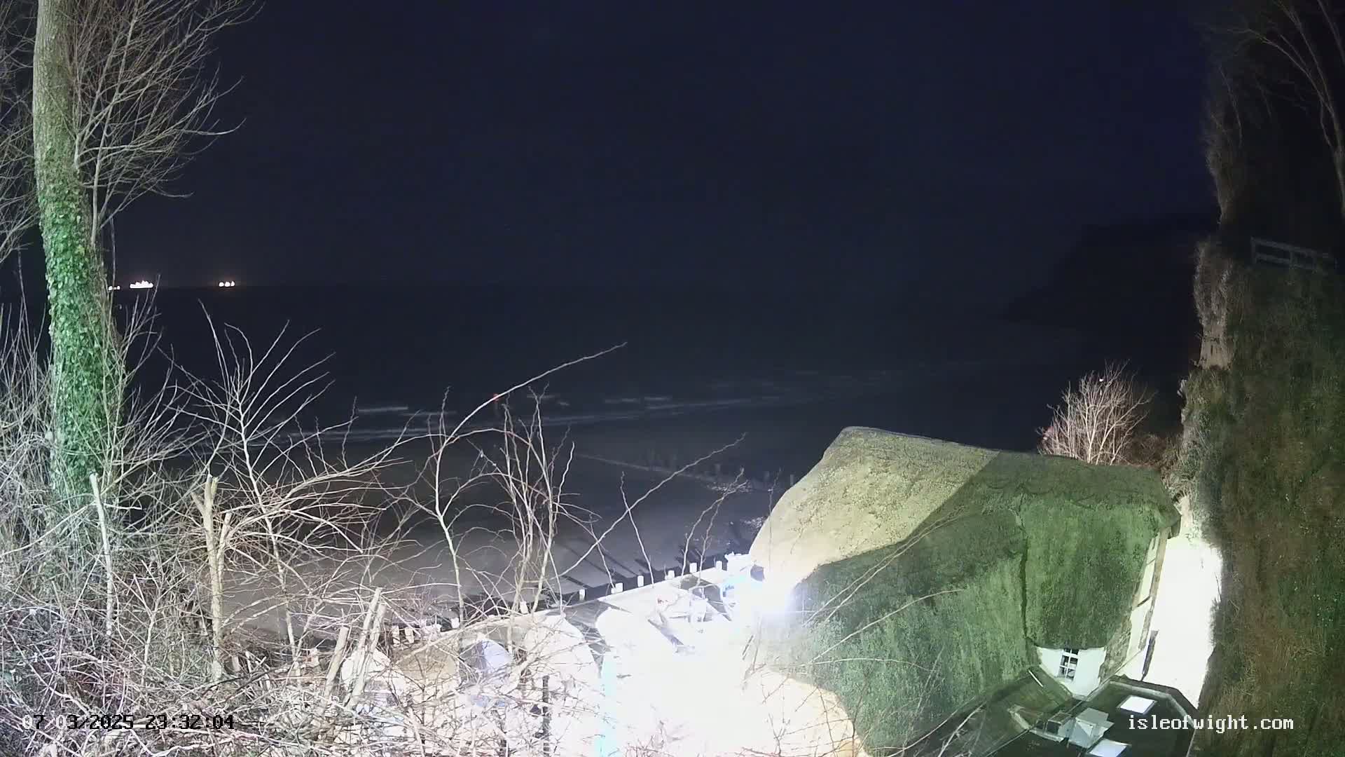 A nighttime view of a dark ocean with small waves, a sandy beach, and a thatched-roof building perched on a cliff overlooking the water.