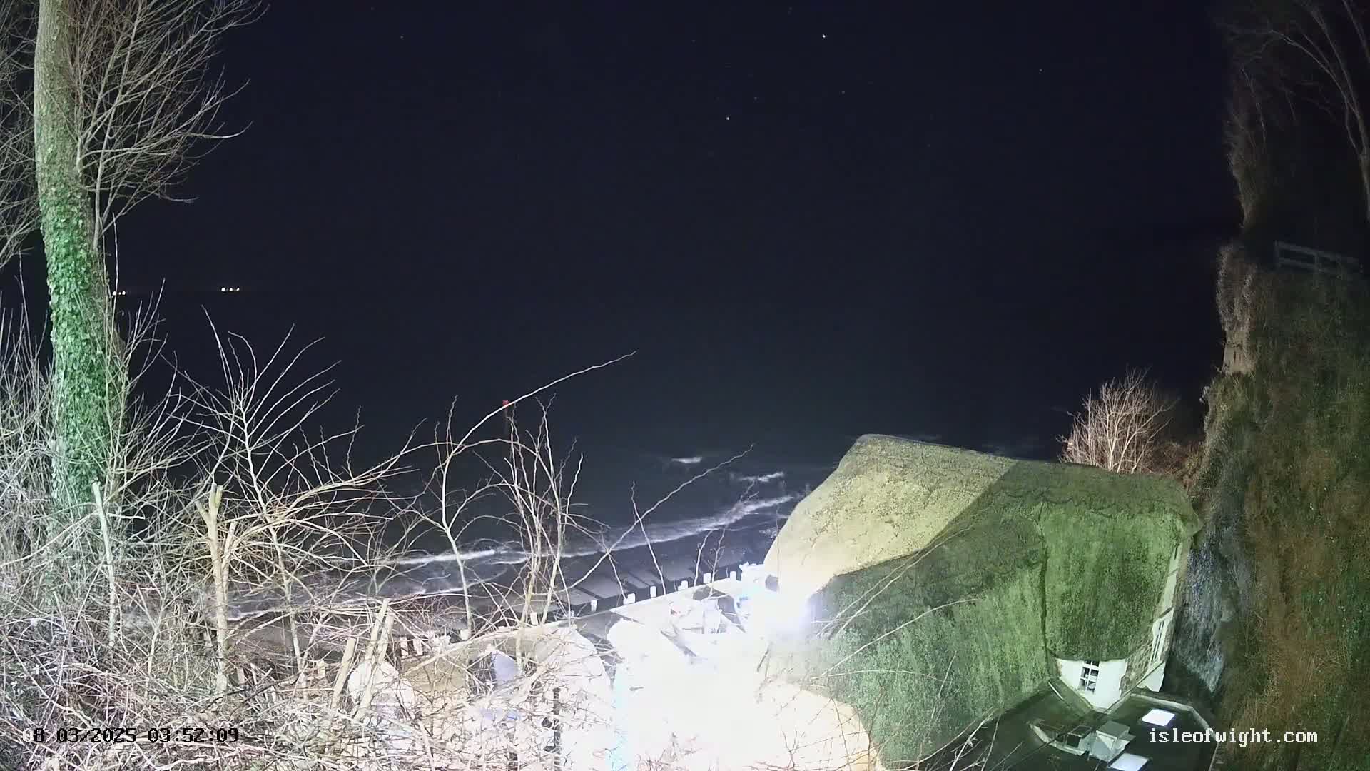 A thatched-roof building sits atop a cliff overlooking a dark, wavy sea at night under a starry sky.