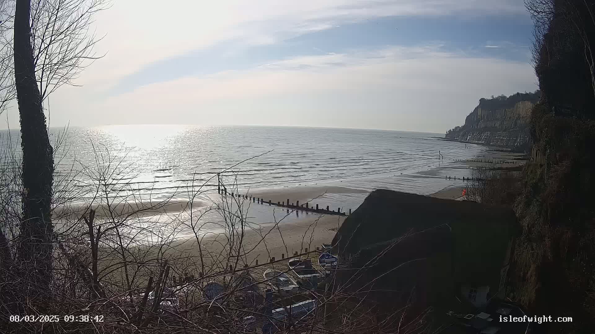 A view from a clifftop shows a calm, bright ocean with a sandy beach, some small boats, and a person in the distance on a sunny day.