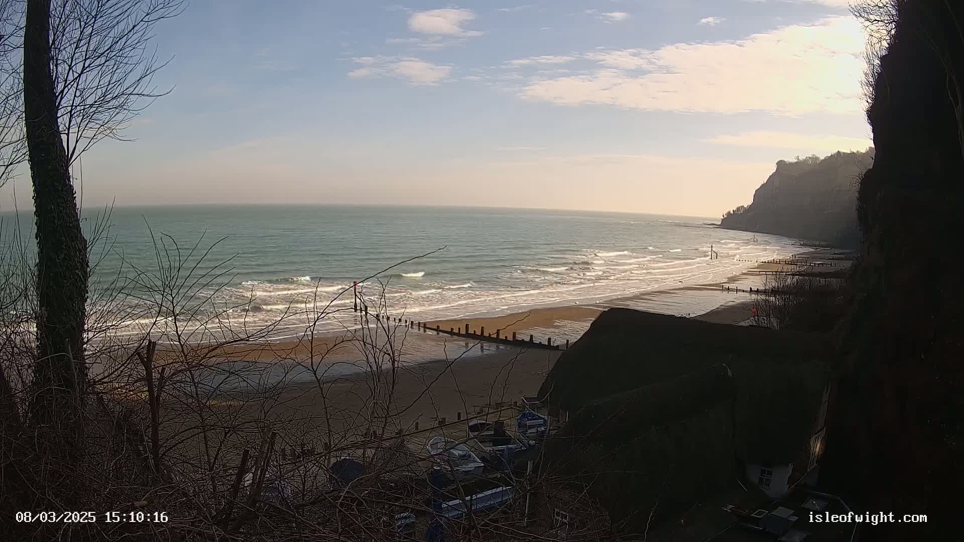 A sandy beach with gentle waves stretches along a coastline under a mostly clear sky, viewed from a clifftop overlooking several boats below.