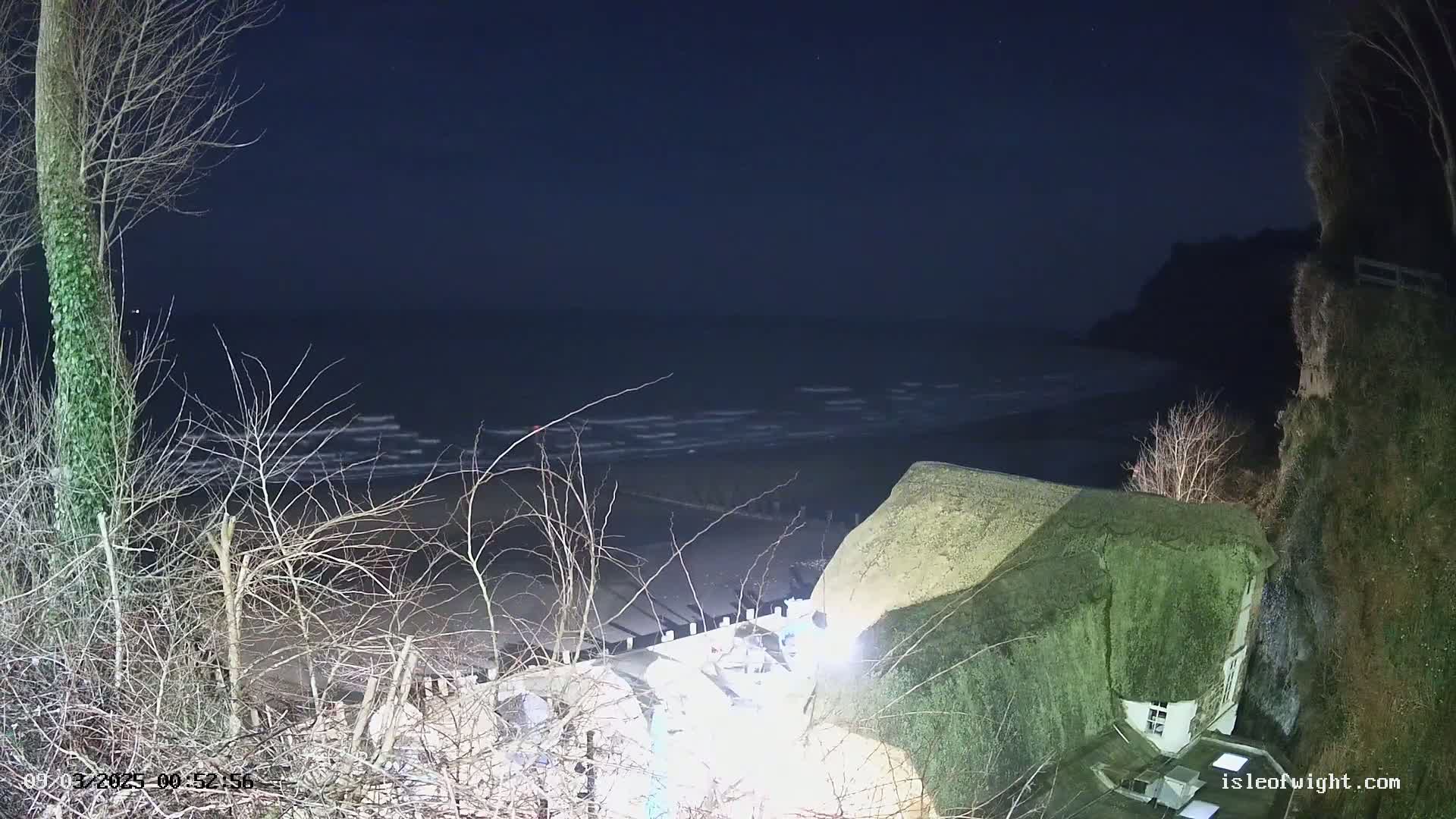 A dark, nighttime scene shows a thatched-roof building perched on a cliff overlooking a wavy ocean under a clear, starless sky.