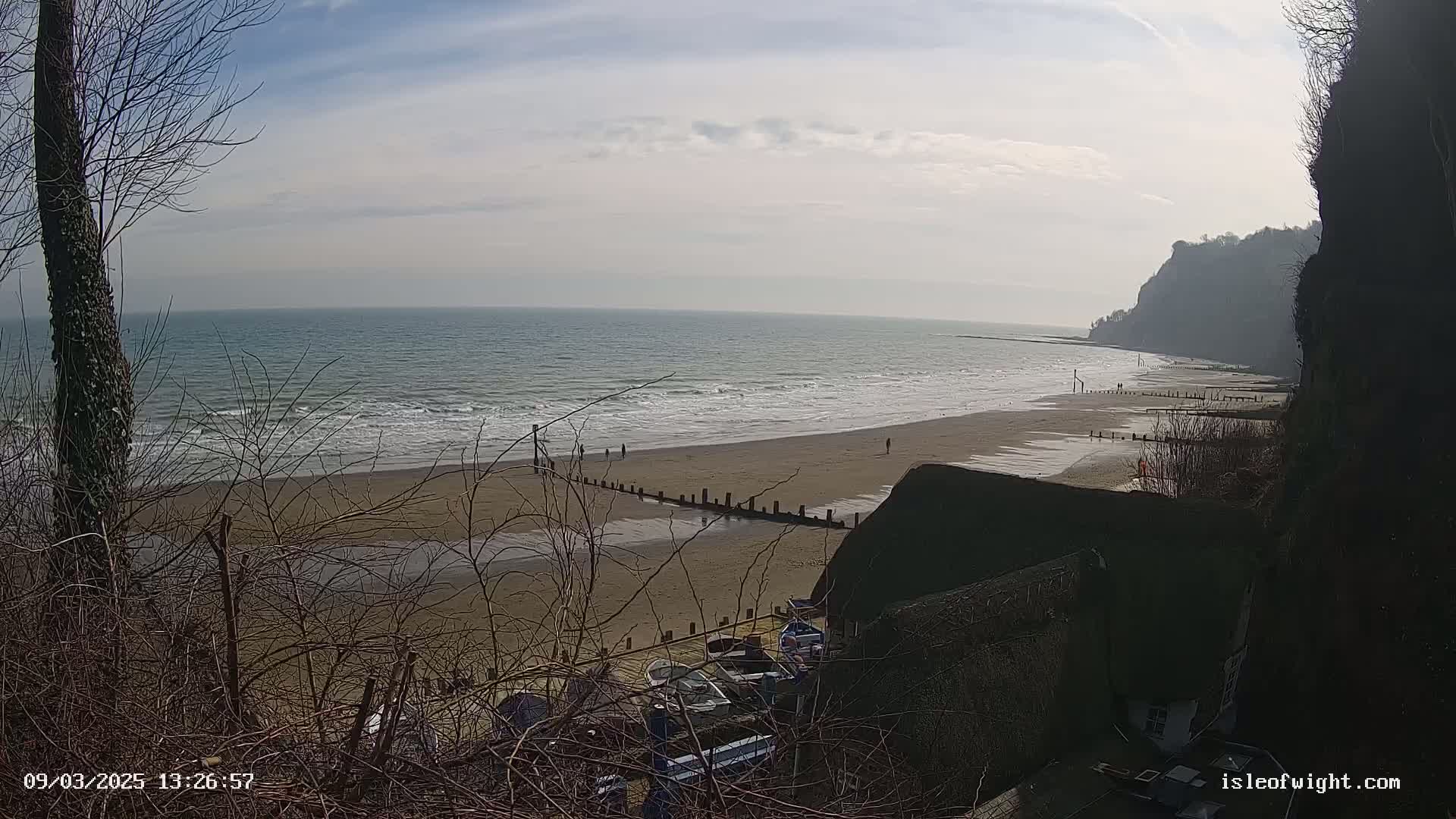 A sandy beach with gentle waves and a few people stretches out below a partially-visible cliff, all under a mostly sunny sky.