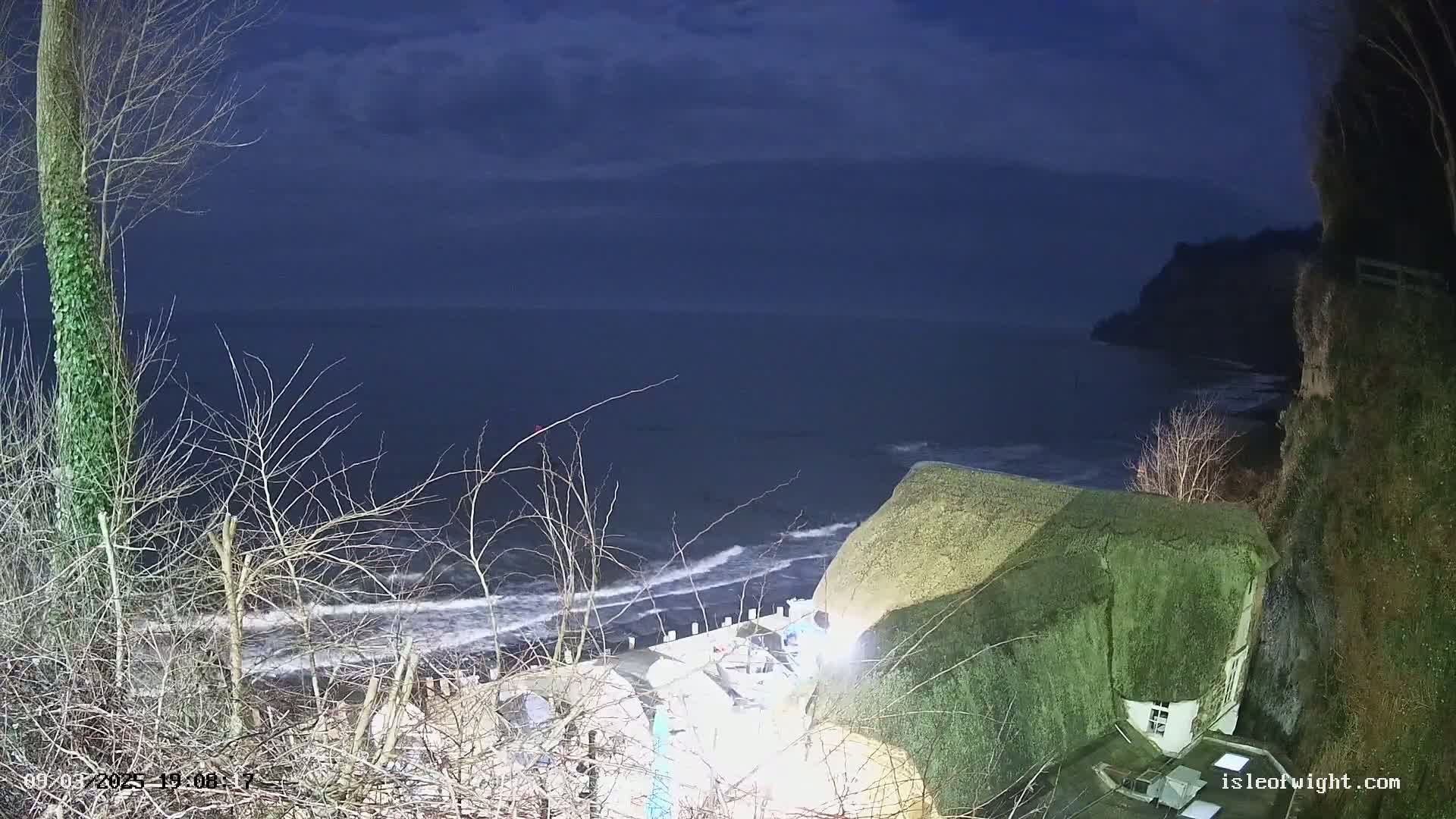 A nighttime coastal scene shows a dark sea with small waves lapping a shore, with a thatched-roof building perched on a cliff overlooking the water under a mostly cloudy sky.