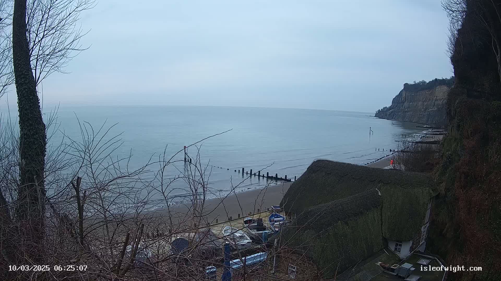 A calm, overcast day reveals a sandy beach, a line of wooden posts in the water, several small boats, and a thatched-roof building nestled into a cliffside overlooking the sea.