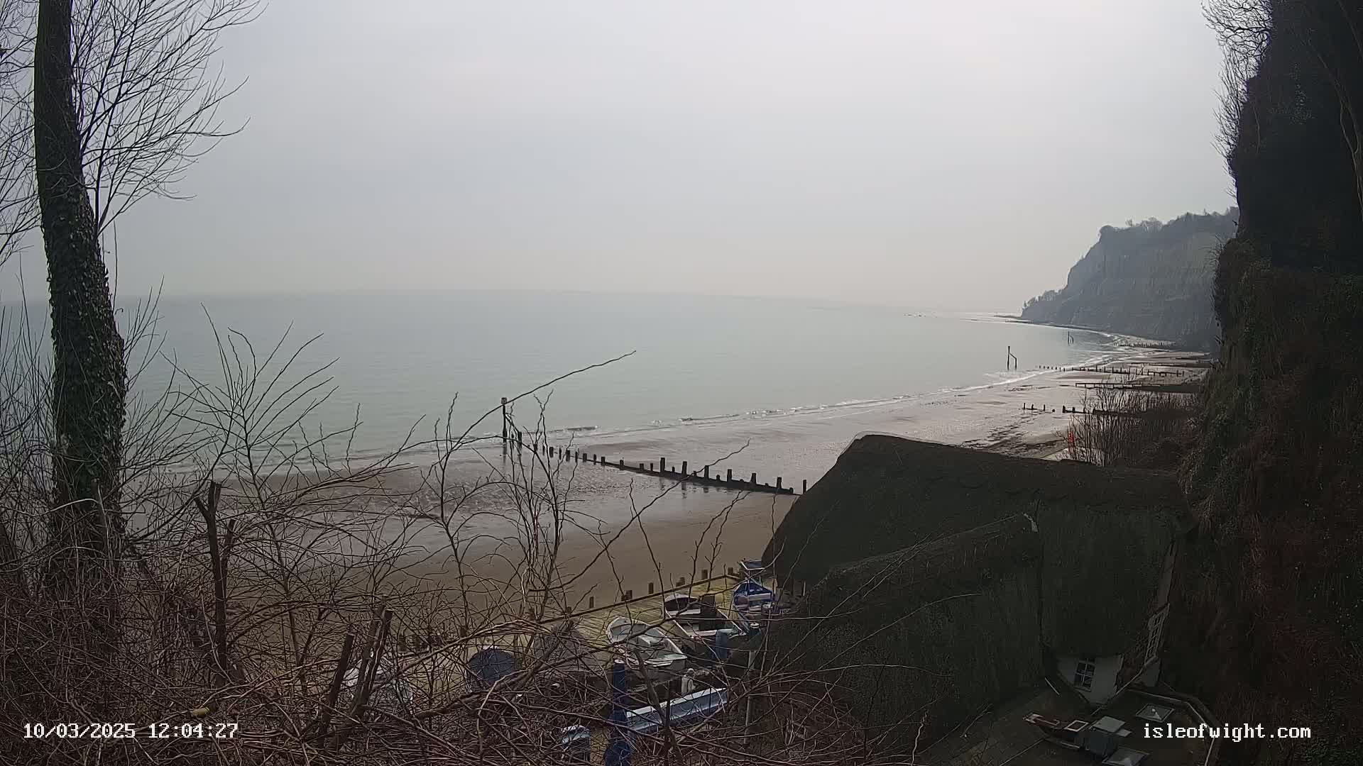 A hazy view from a clifftop shows a beach with several small boats pulled ashore near a thatched-roof building, with a calm sea and distant cliffs under a gray sky.