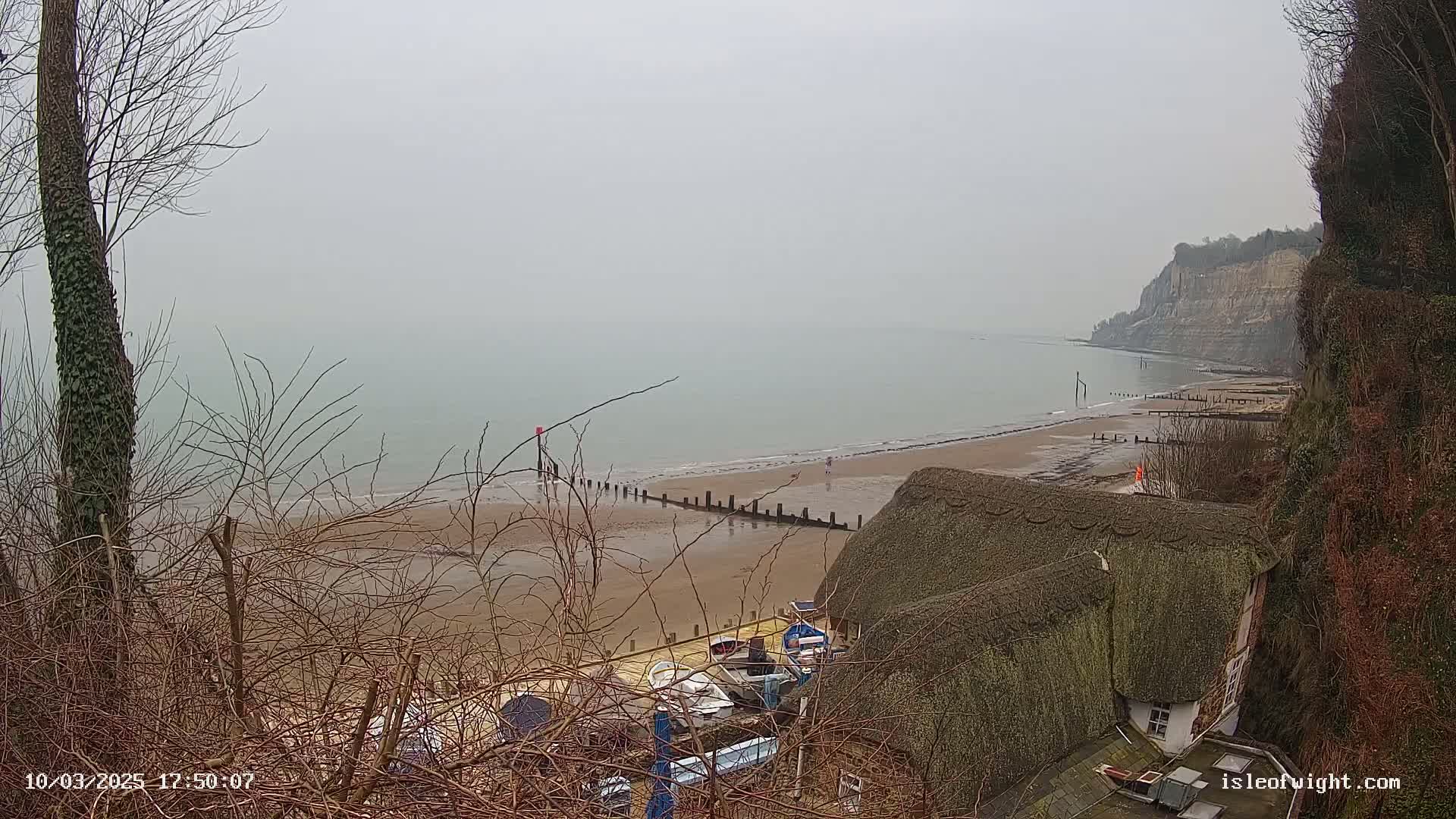 A foggy view of a sandy beach with a few people, wooden posts in the water, boats pulled ashore, and a thatched-roof house situated on a cliff overlooking the sea.