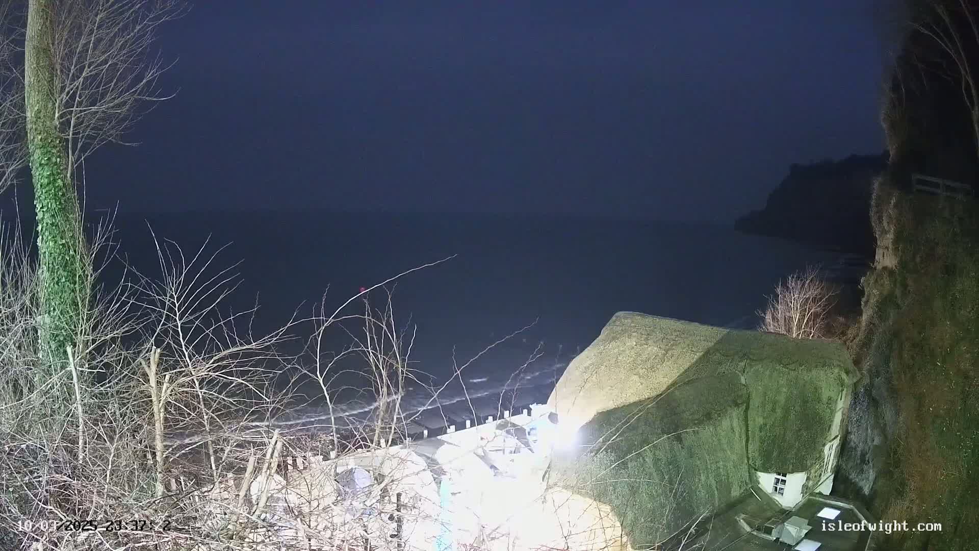 A dark, nighttime coastal scene shows a thatched-roof house perched on a cliff overlooking a dark sea with gentle waves under a mostly clear, dark sky.