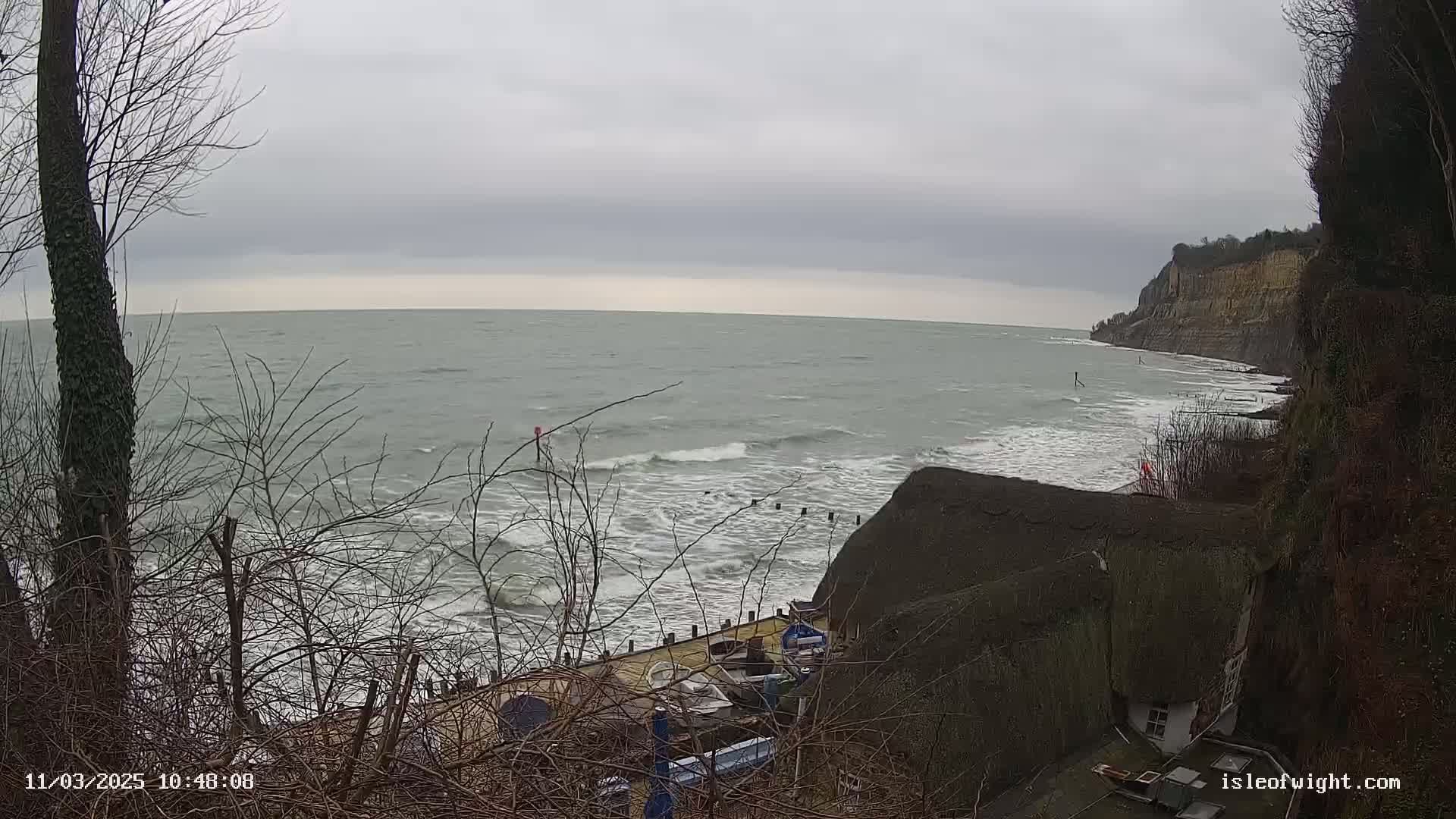 A gray, overcast sky hangs over a choppy sea crashing against a rocky shoreline where several small boats are moored near a thatched-roof house nestled into the cliffside.