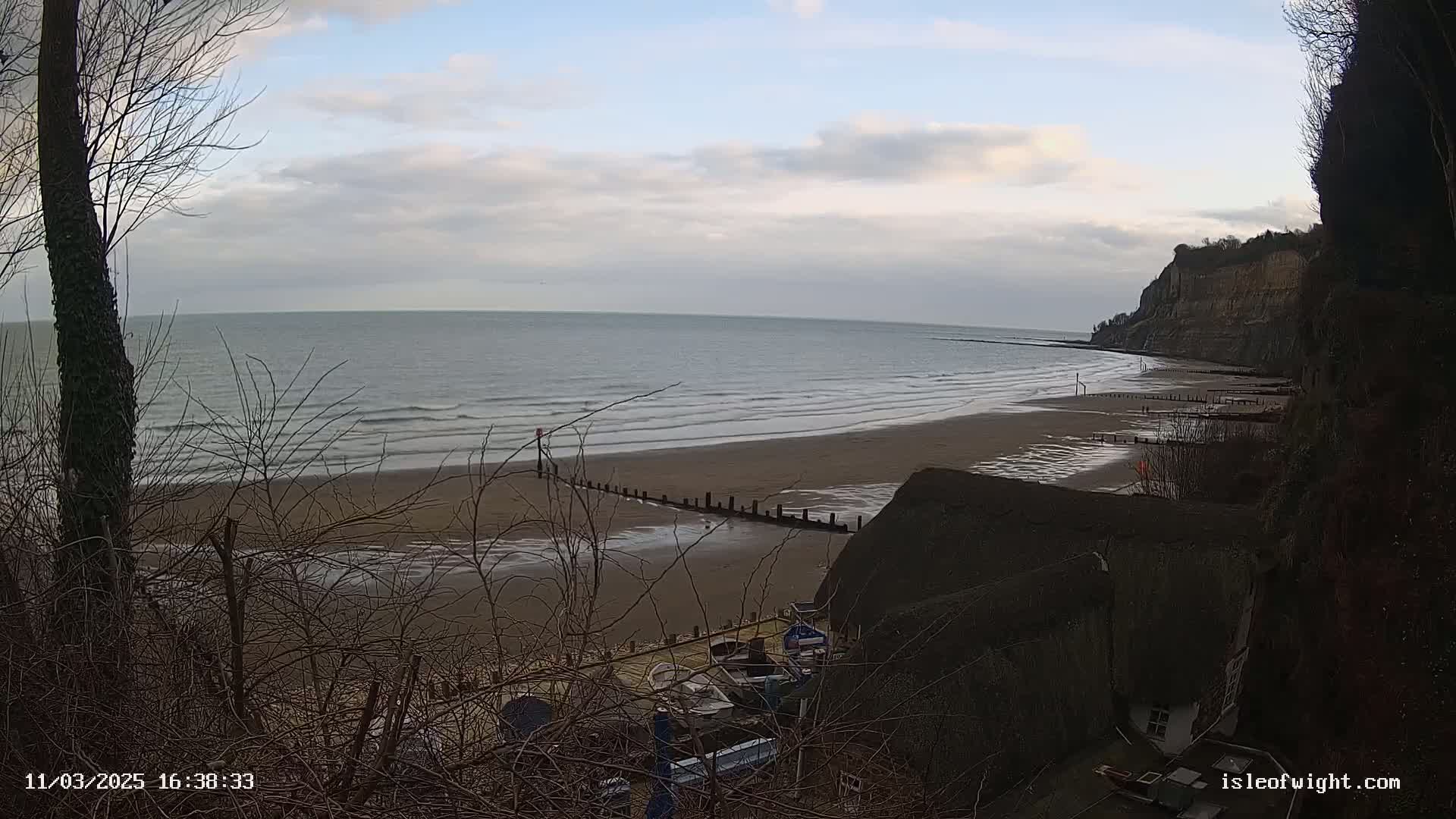 A sandy beach with gentle waves under a cloudy sky is viewed from a clifftop, overlooking a small harbor with boats.