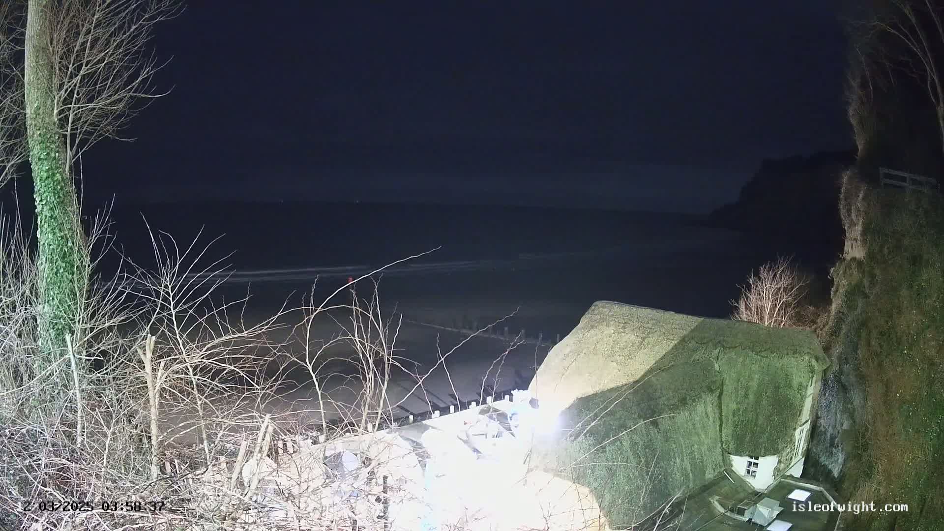 A nighttime, seaside view shows a thatched-roof building perched on a cliff overlooking a dark ocean and beach.