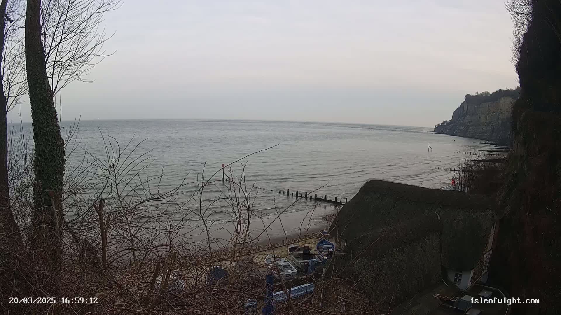 A calm, overcast day reveals a view of a beach with several small boats, a thatched roof building, and a cliff face extending into a body of water.