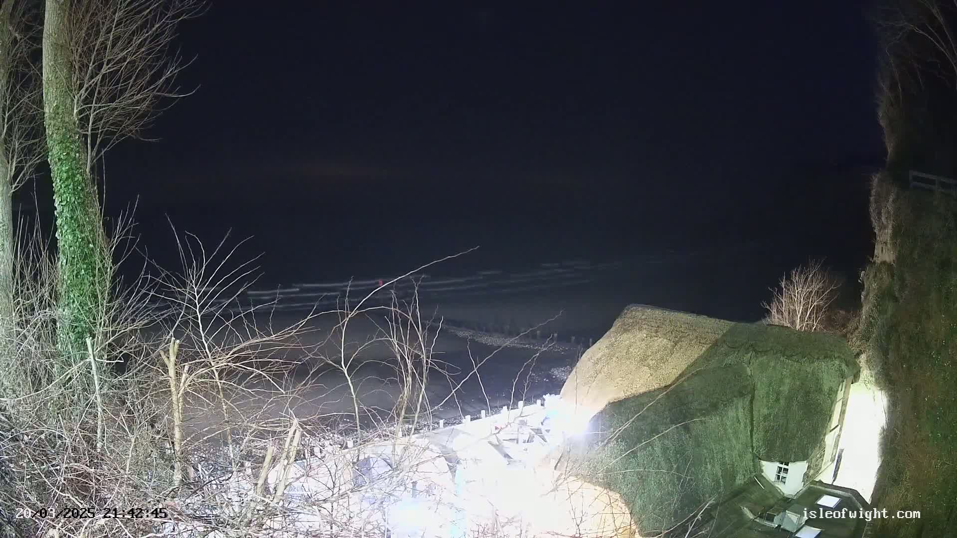 A thatched-roof house sits atop a cliff overlooking a dark, wavy ocean at night.
