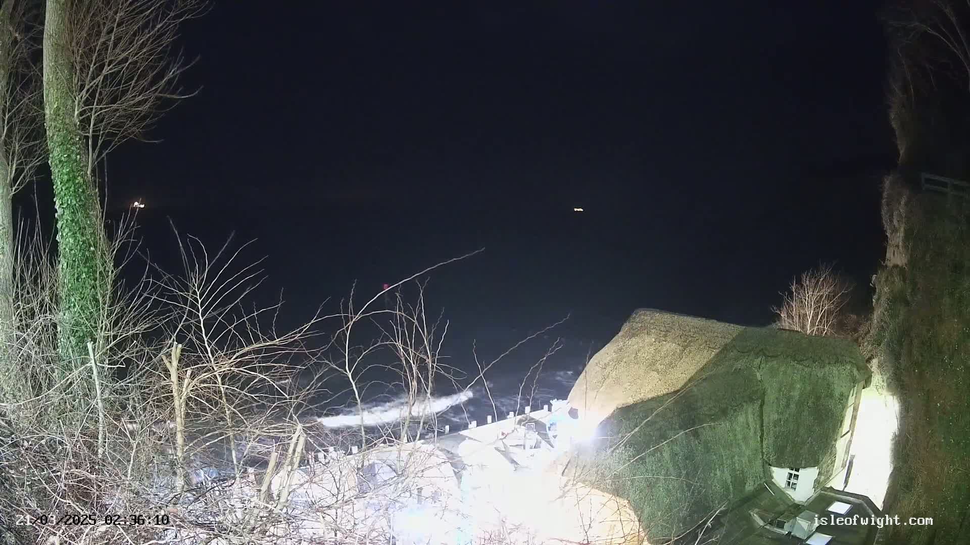 At night, a thatch-roofed house sits precariously close to a dark, wavy sea under a clear, dark sky.
