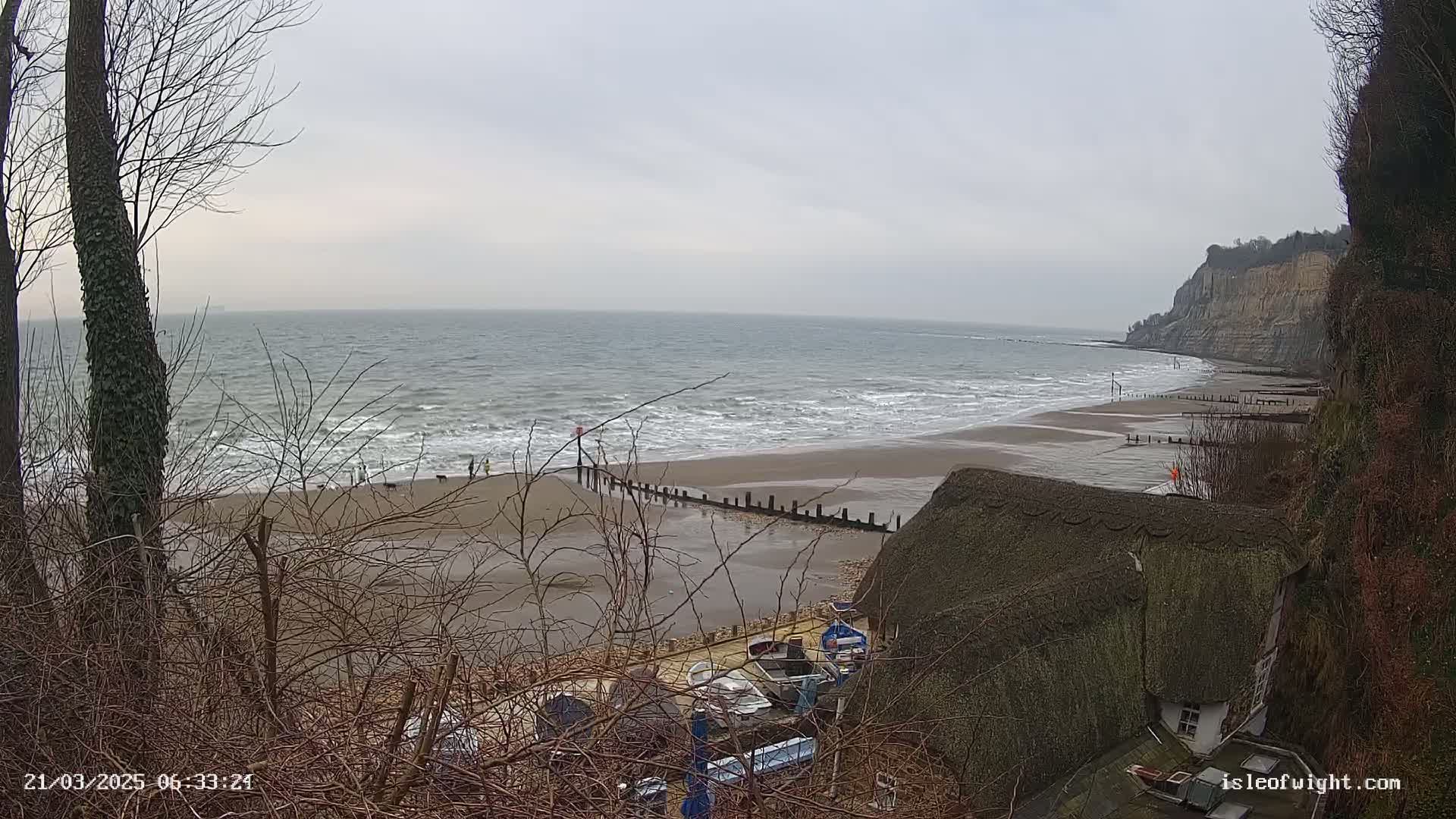 A sandy beach with small waves under an overcast sky is viewed from a hillside, showing a thatched-roof house and a few boats in the foreground.