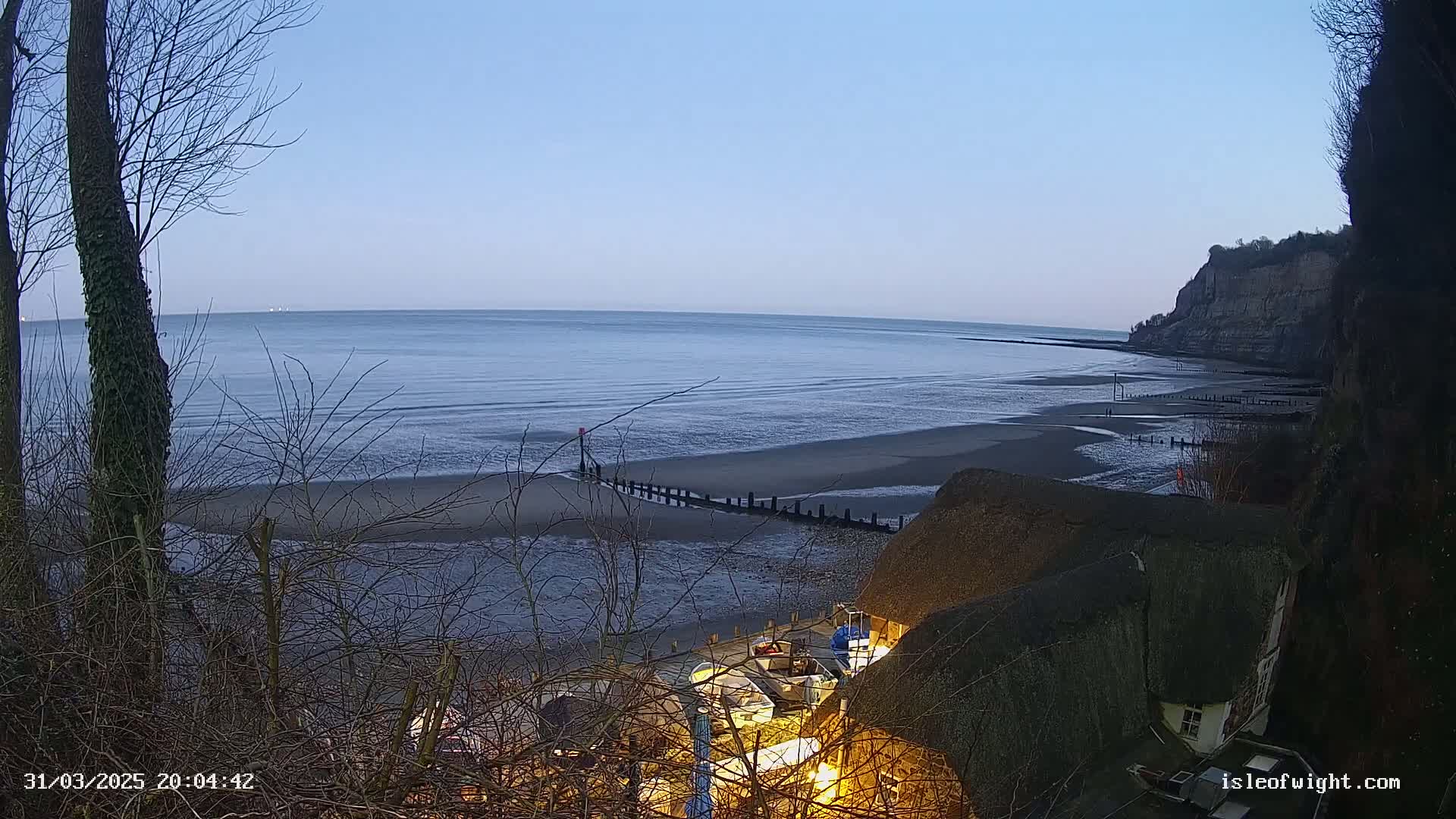 A calm, twilight sea stretches to the horizon beside a sandy beach, with a few small boats visible near a thatched-roof building nestled at the base of a cliff.