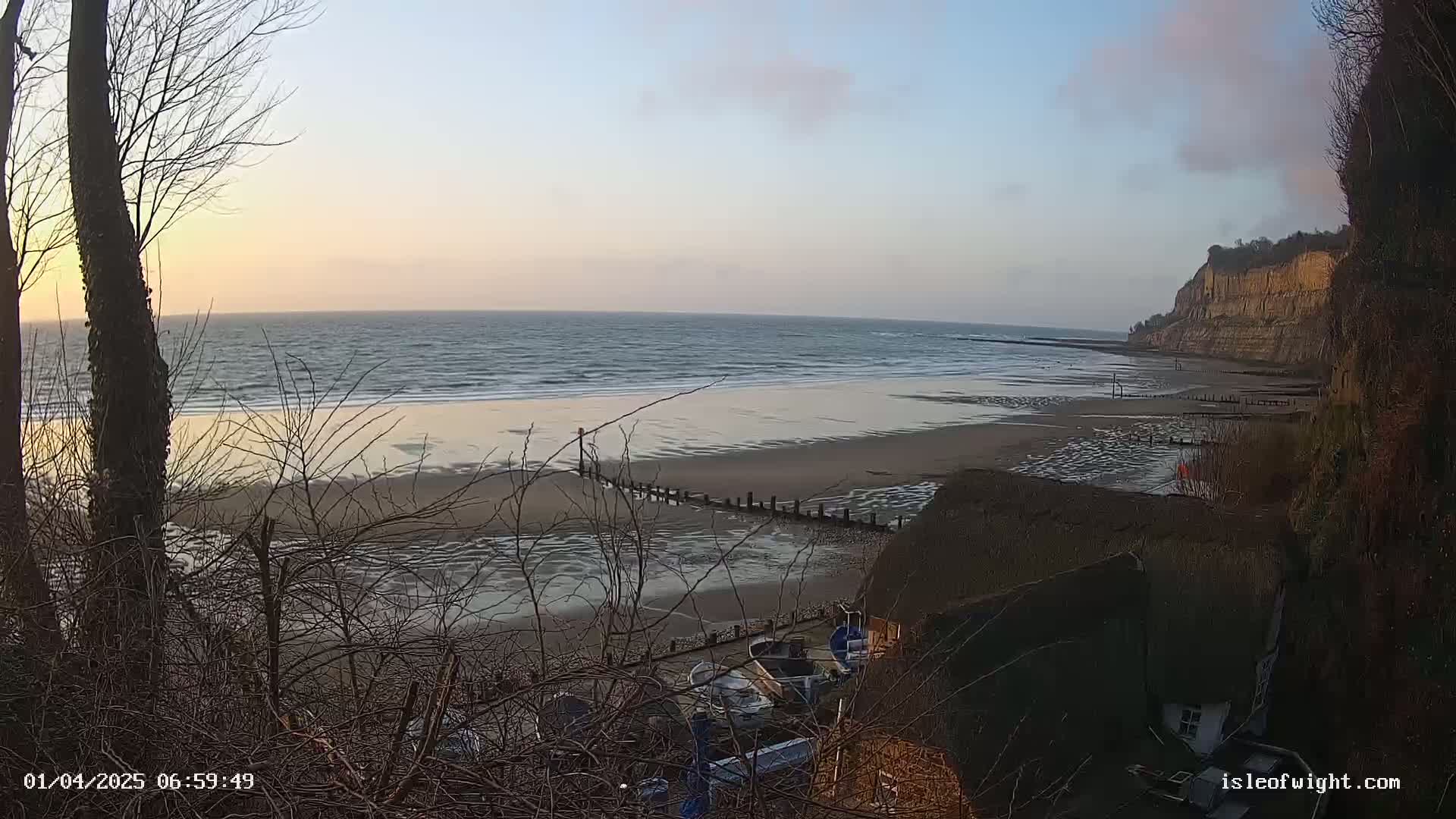 A sandy beach with gentle waves at sunset is seen from a high vantage point, with several small boats in a cove below and a tall cliff in the background.