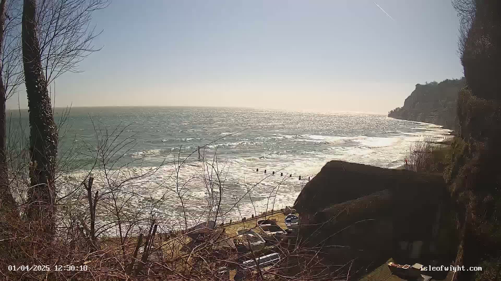 A view from a cliff overlooking a wavy sea on a bright, sunny day, with several boats parked below and bare winter trees in the foreground.