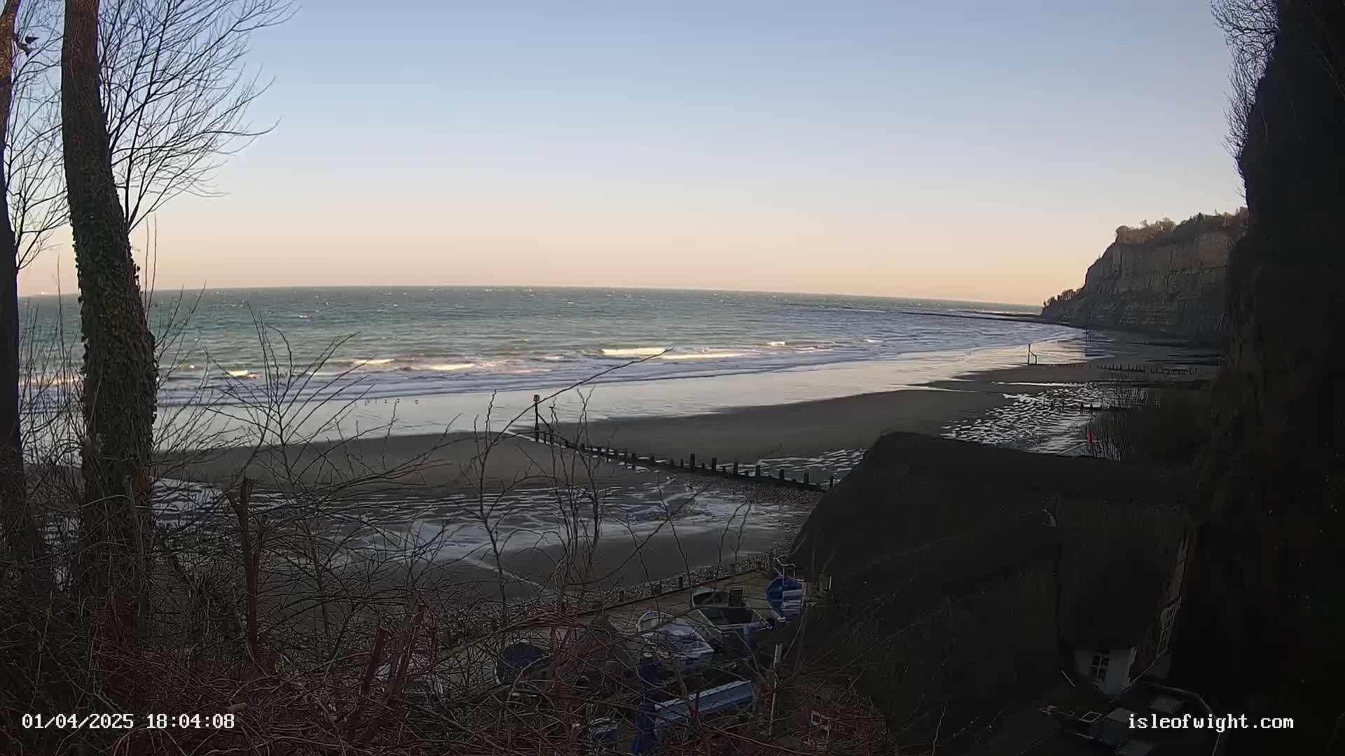 A view from a clifftop shows a windy, wavy ocean meeting a wide sandy beach with some boats pulled ashore below, partially obscured by vegetation.