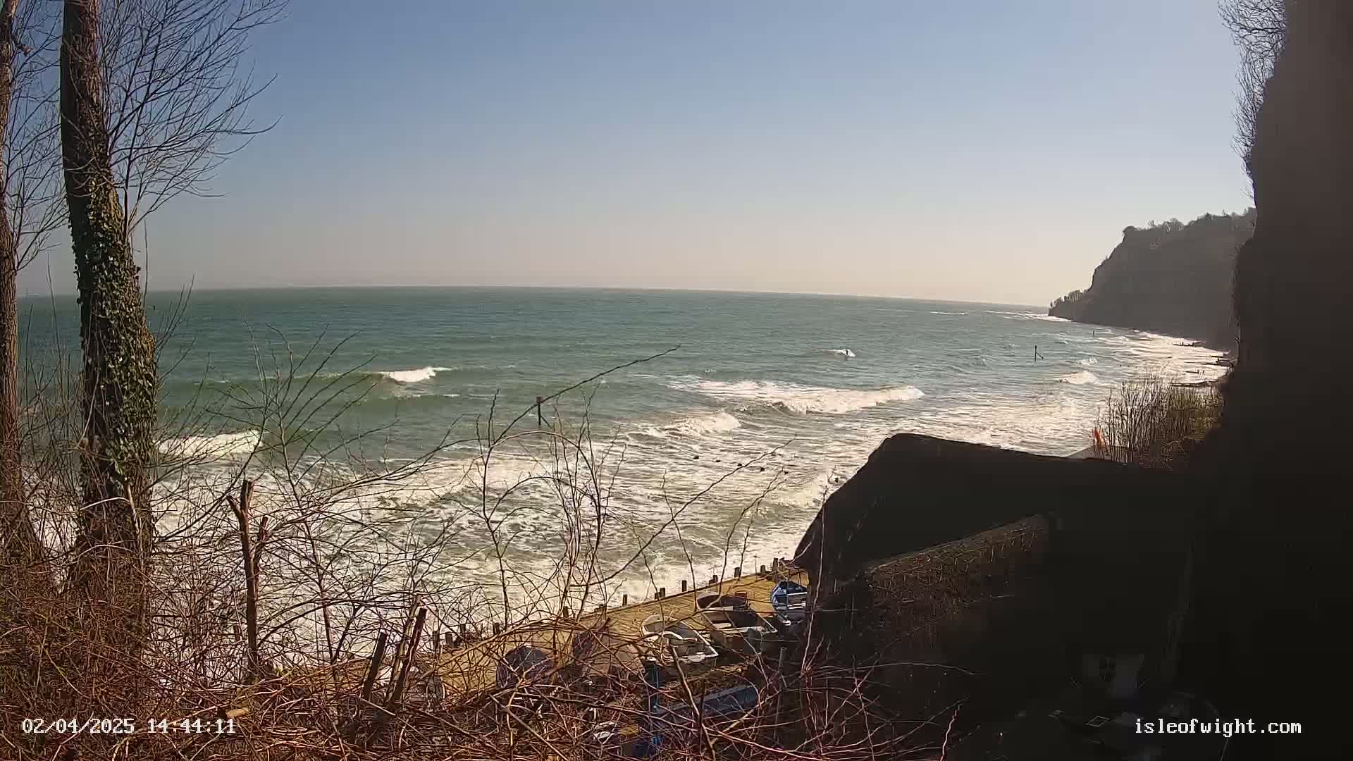 A view from a clifftop shows a rocky shore and choppy ocean waves under a clear blue sky.