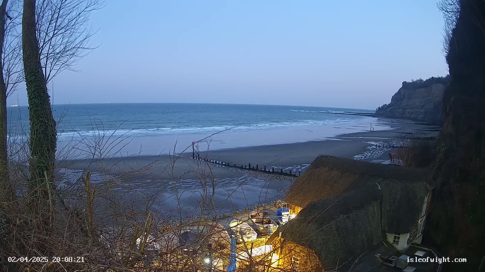 A tranquil beach scene at dusk shows a calm ocean with small waves lapping a sandy shore, backed by a cliff, and partially obscured by a thatched roof building and bare winter trees.
