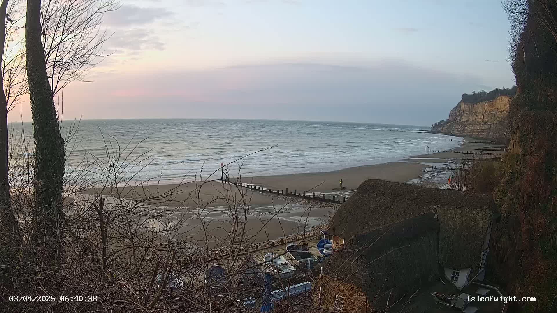 A sandy beach with gentle waves under a pale sunrise sky is viewed from a clifftop, showing several small boats near a thatched roof building.