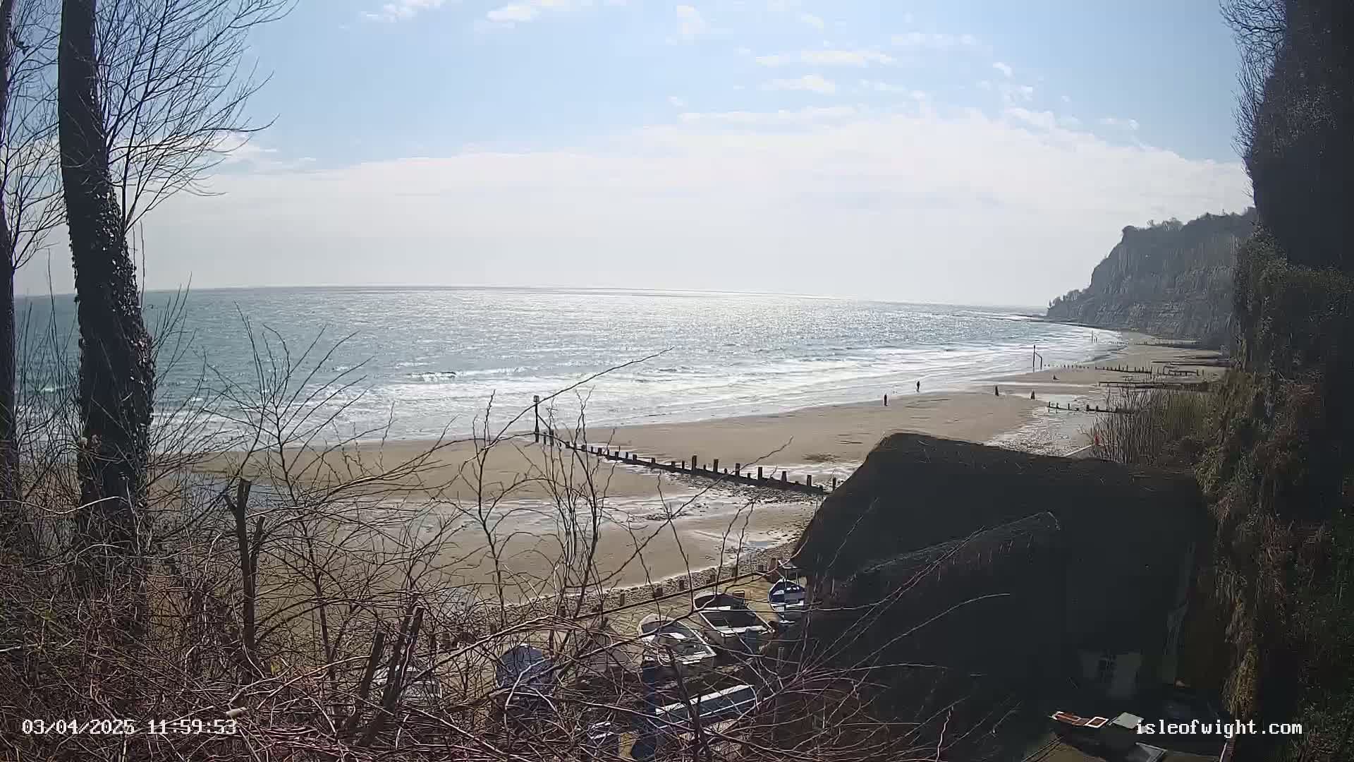 A sandy beach with gentle waves under a partly cloudy sky is seen from a high vantage point, with a few people visible on the beach and several small boats at the water's edge near a thatched roof building.