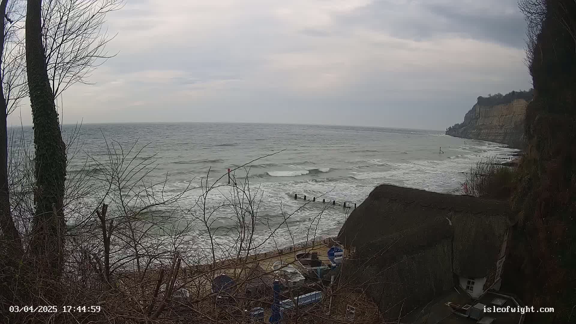 A view from a cliff overlooking a choppy sea with waves breaking on a rocky shore, a thatched-roof house nestled below, and bare winter trees in the foreground under a cloudy sky.