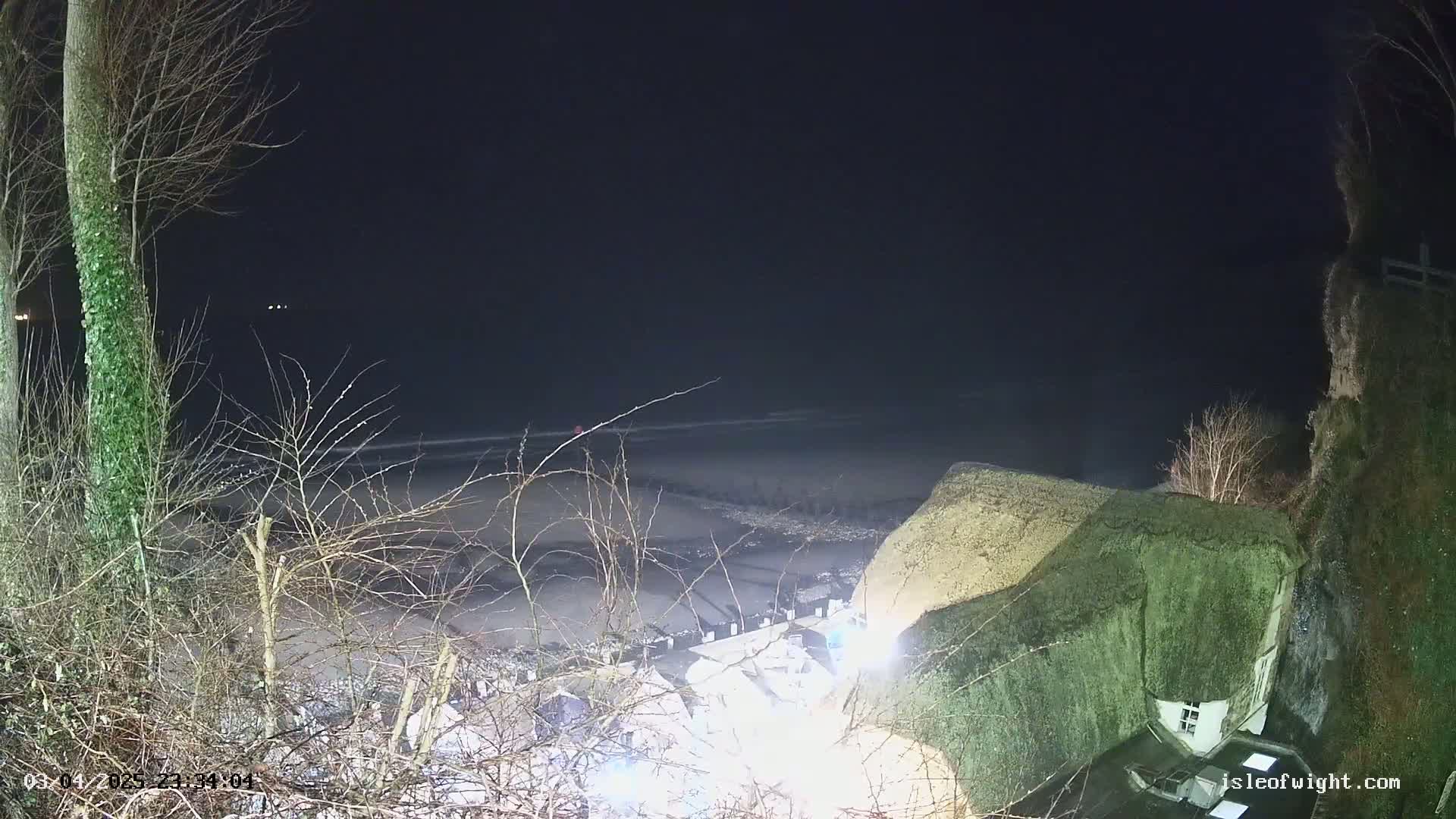 A nighttime view shows a dark beach and ocean under a clear, dark sky, with a thatched-roof house partially visible on a cliff overlooking the beach.