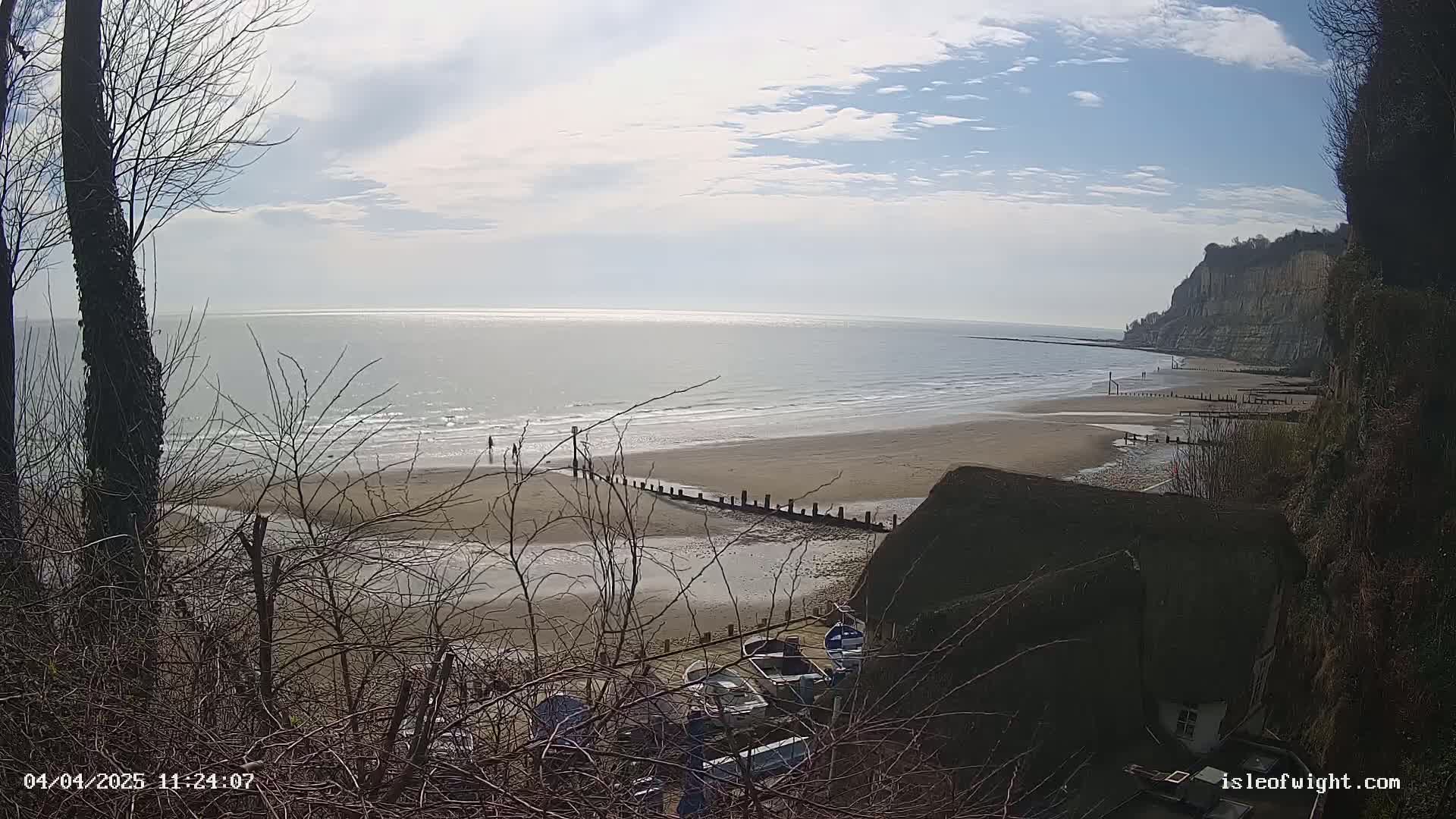 A sandy beach stretches below a steep cliff under a partly sunny sky, with several boats visible near a thatched-roof building and bare winter trees in the foreground.