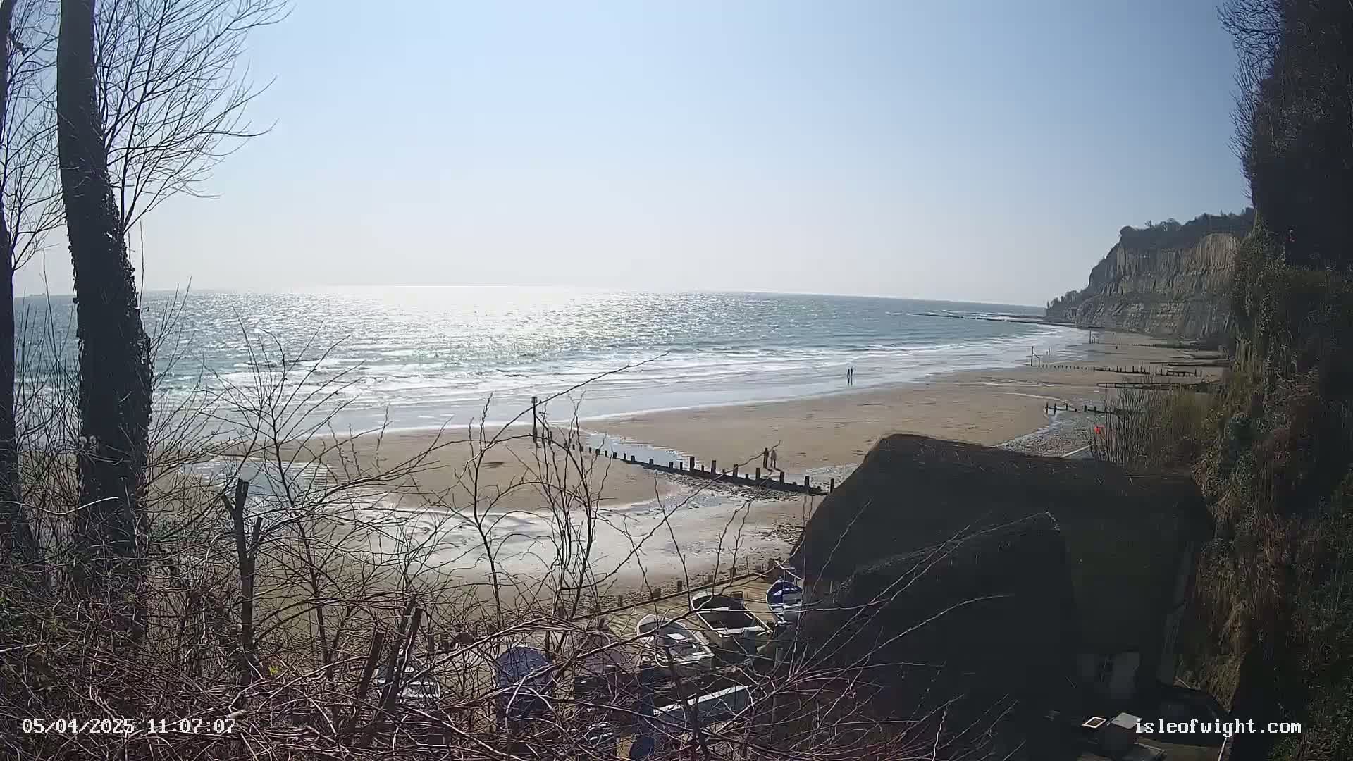 A sandy beach with a calm, sunlit sea stretches out beneath a bright sky, viewed from behind leafless bushes overlooking a small collection of boats near the cliff edge.