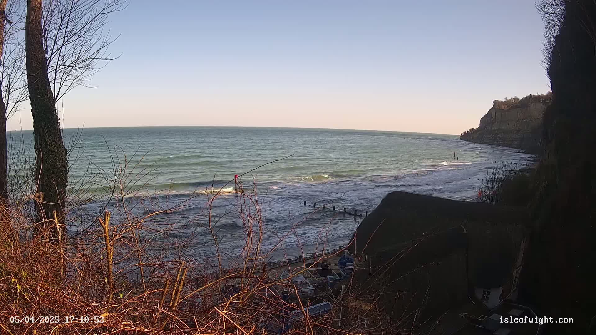 A view from a clifftop shows a calm sea with small waves lapping a beach below, under a clear, bright sky.