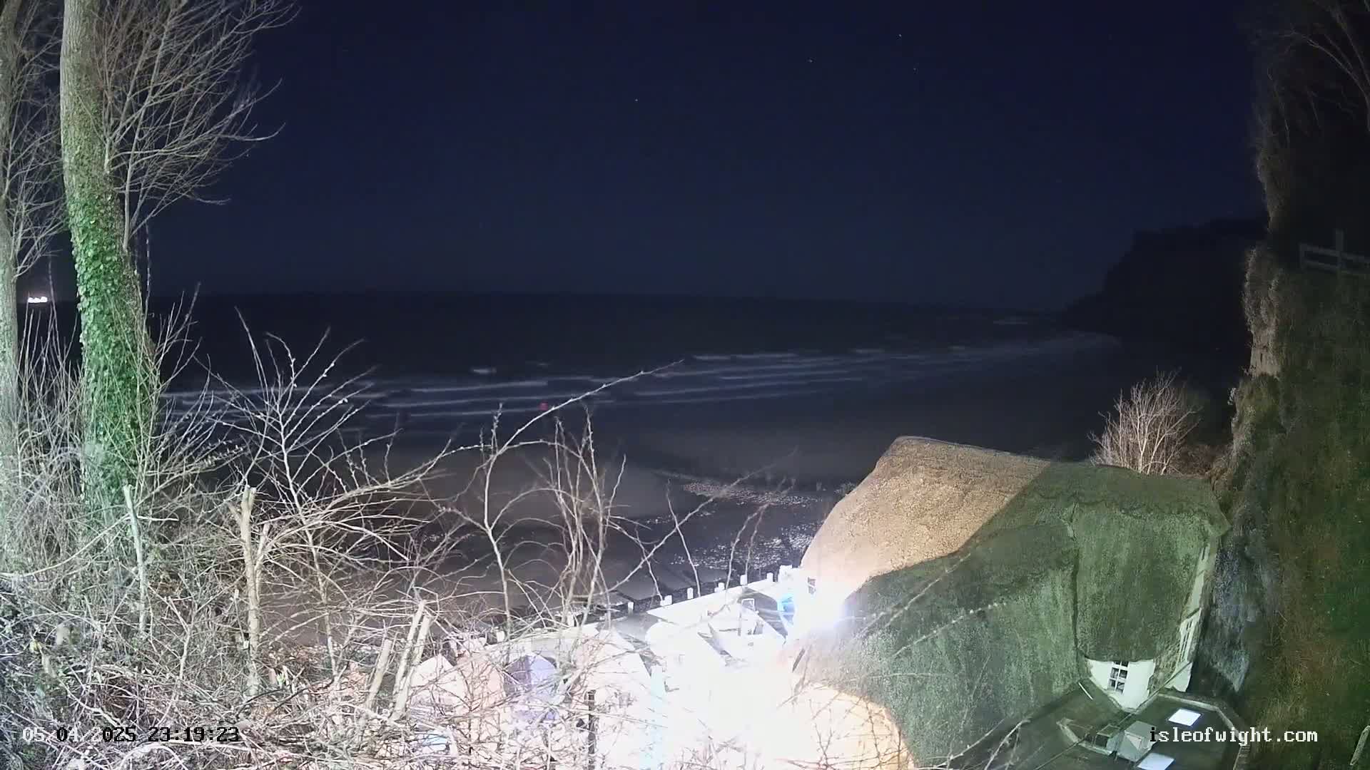 A nighttime view of a dark, wavy ocean under a clear, star-studded sky, with a thatched-roof house partially illuminated on a cliff overlooking the beach.