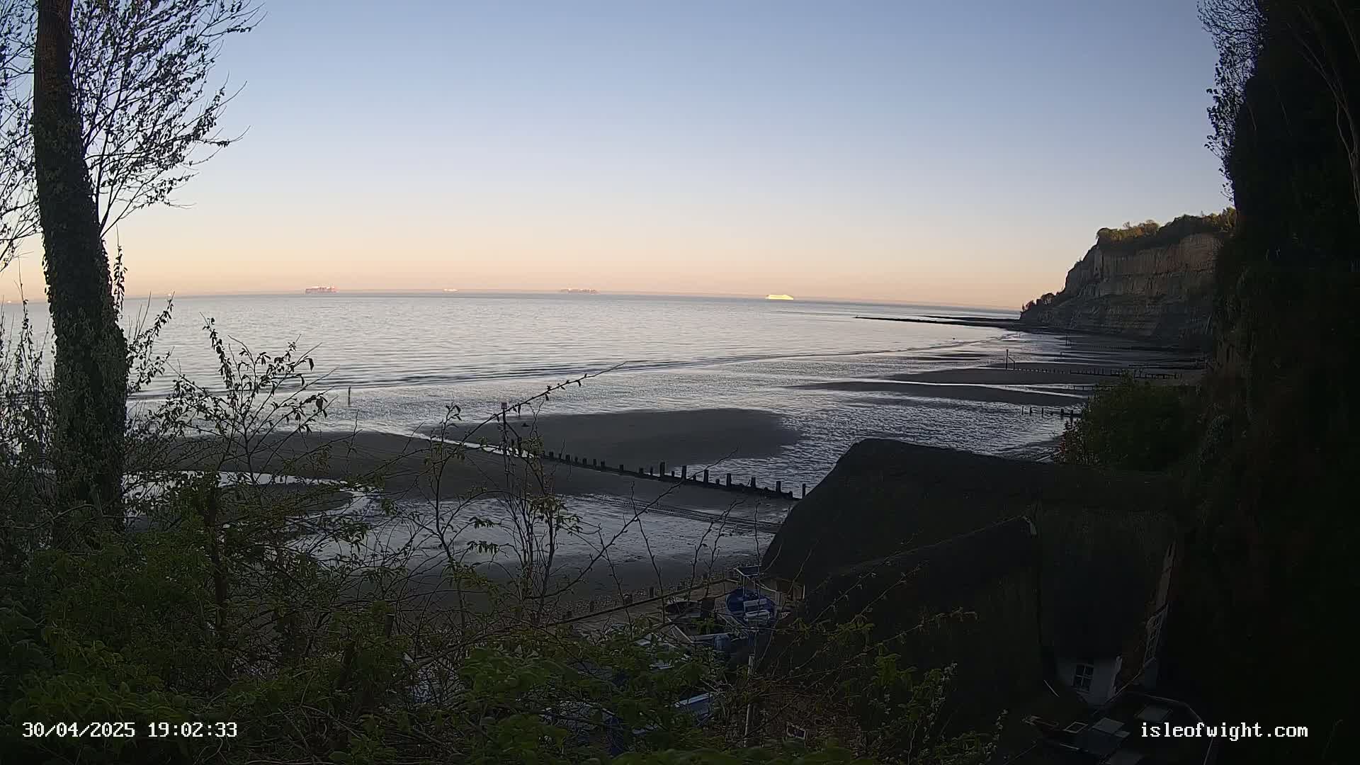 A tranquil beach at sunset, with calm waters, a few boats pulled ashore, and a cliff rising in the background.