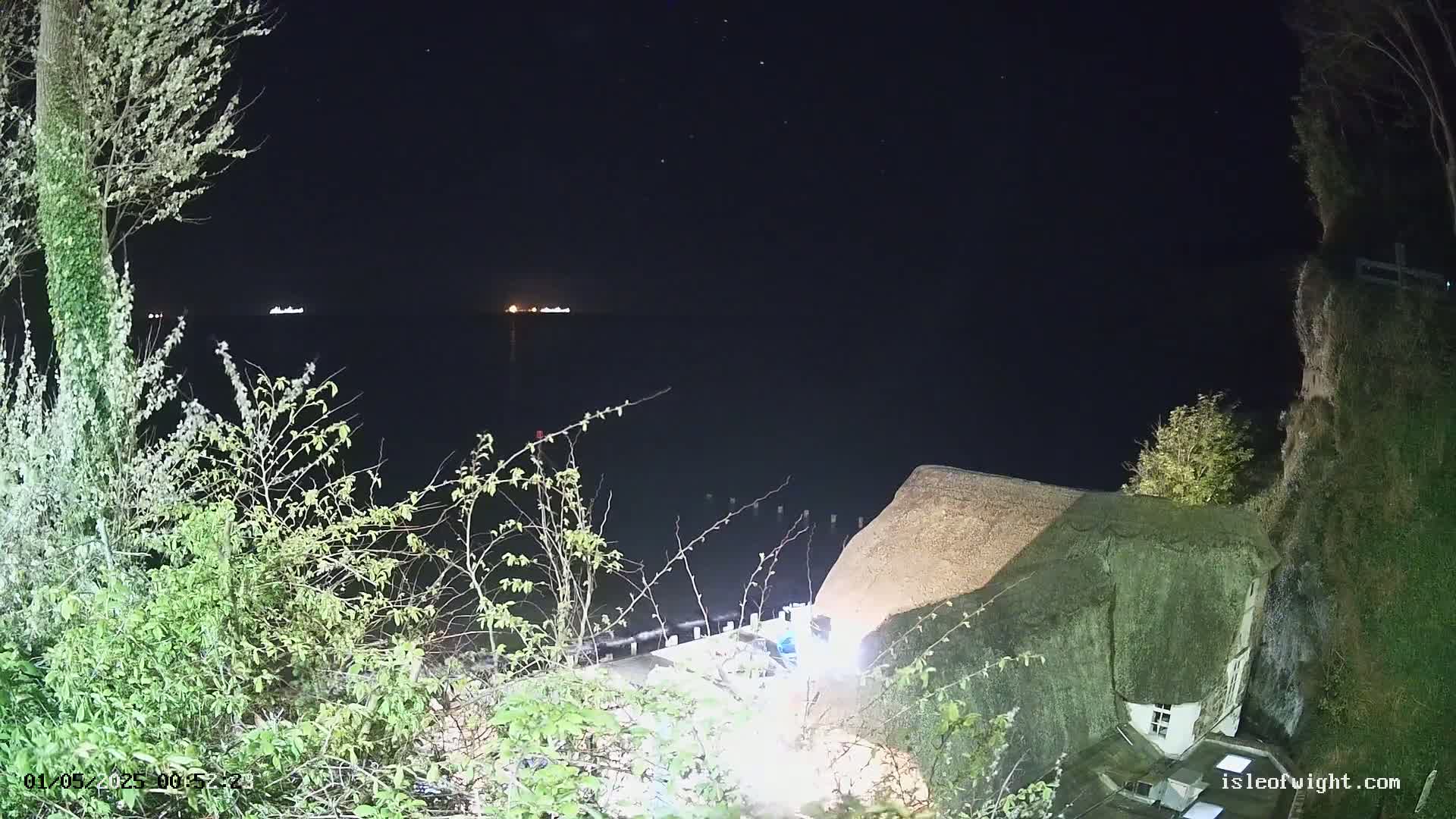 At night, a view from a clifftop shows a dark body of water with distant lights,  vegetation in the foreground, and a partially visible thatched-roof building.