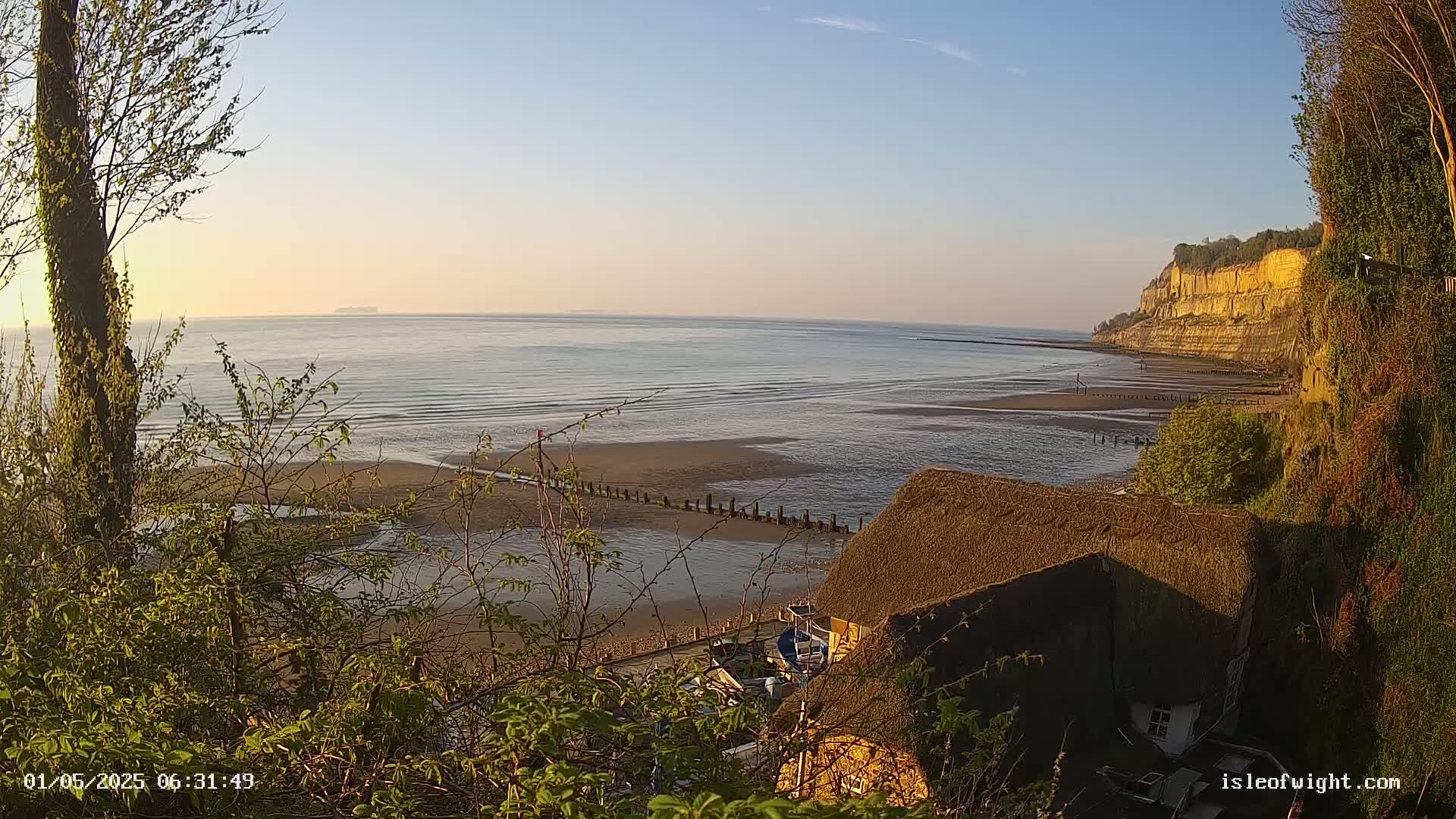 A calm, sunlit ocean scene shows a sandy beach at low tide, a thatched-roof house, and a high cliff in the background.
