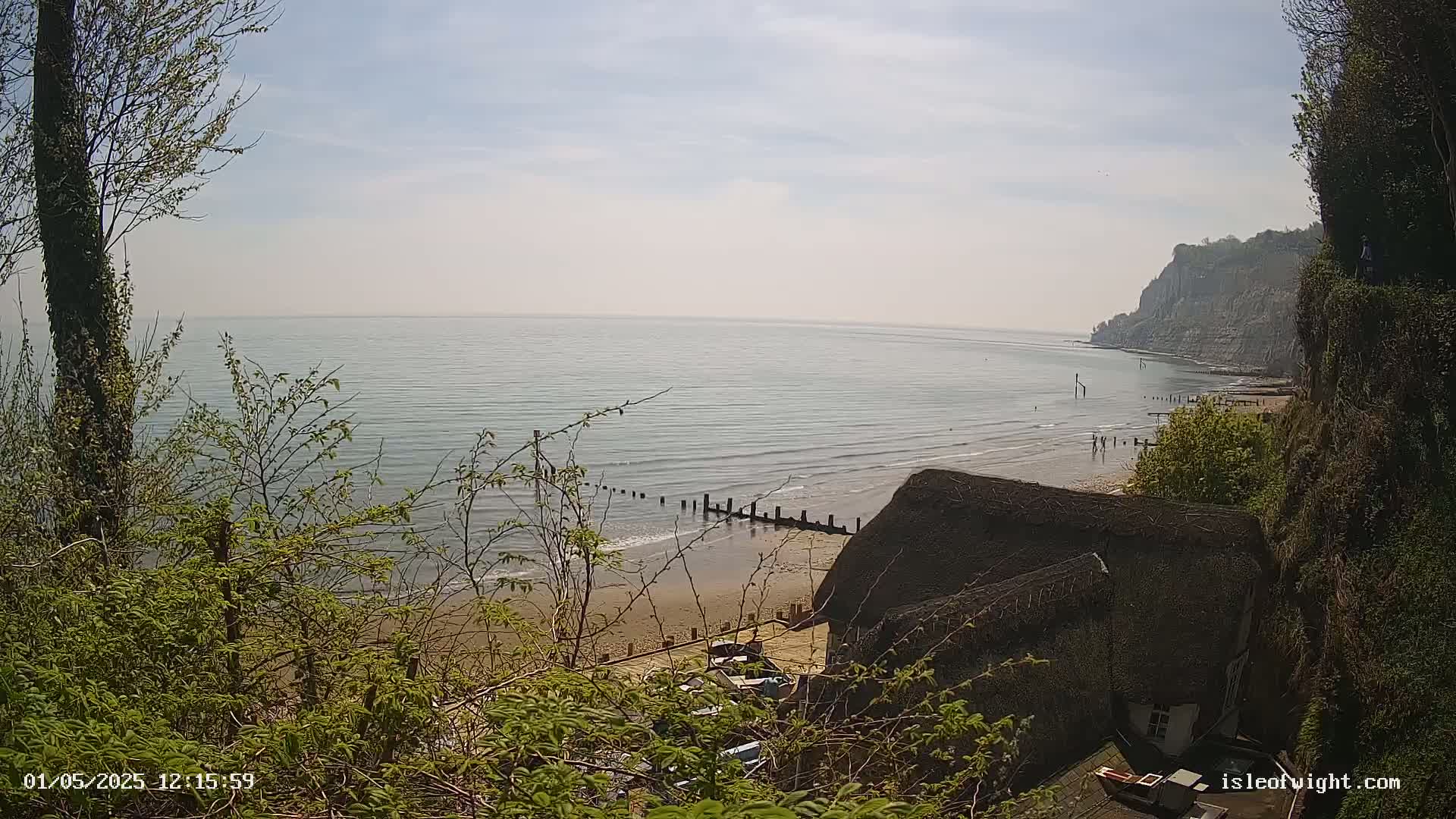 A view from a clifftop shows a calm, light-blue sea gently lapping a sandy beach below, where a thatched-roof building sits near the water's edge, all under a bright, sunny sky.