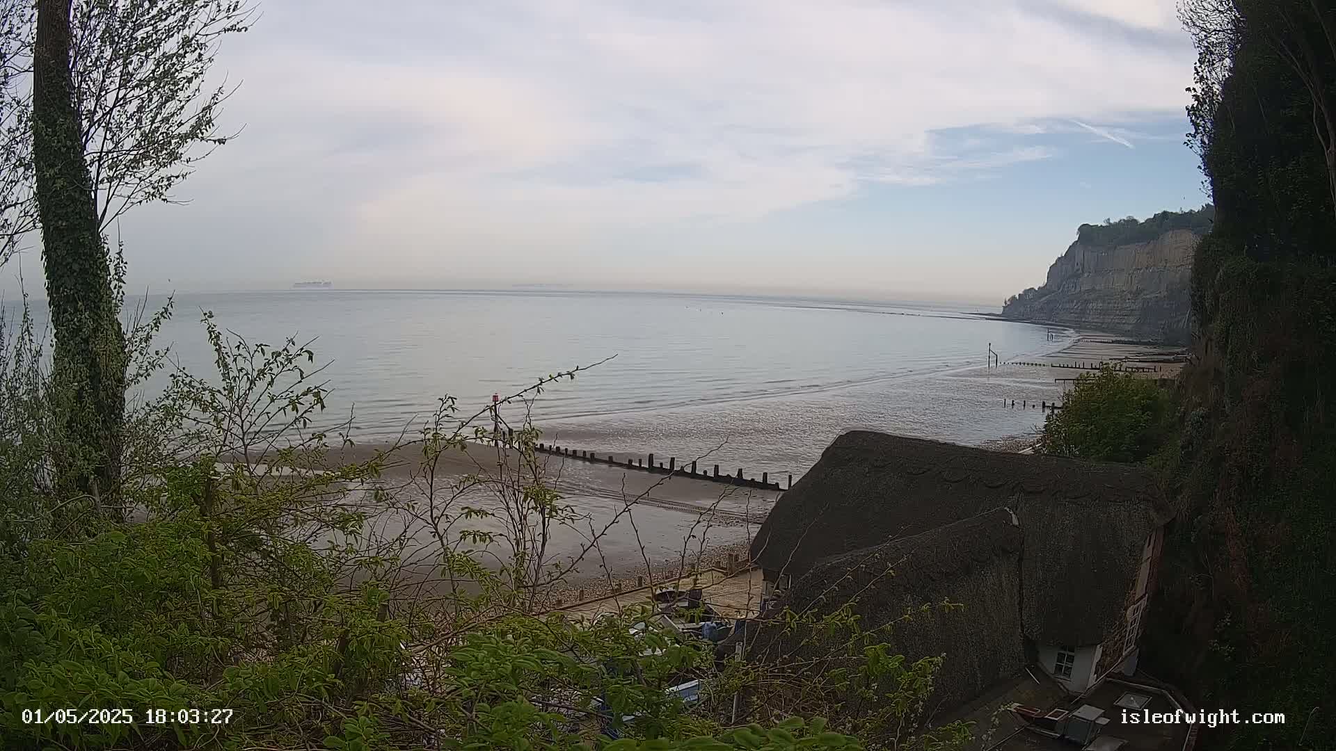 A view from a clifftop shows a calm, pale-grey sea stretching to a hazy horizon, a sandy beach with wooden posts, and a thatched-roof cottage nestled at the base of the cliff.