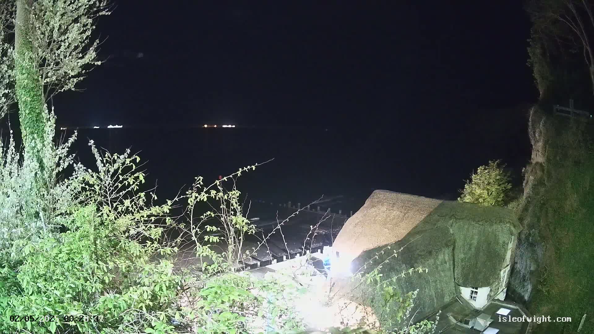 A dark, nighttime coastal scene shows a thatched-roof building partially illuminated, situated on a cliff overlooking a calm, dark body of water with distant lights.