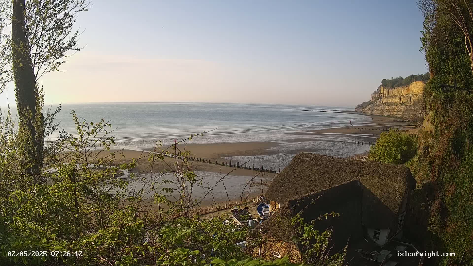 A calm, clear day reveals a sandy beach at low tide, bordered by a thatch-roofed cottage nestled at the base of a tall cliff.
