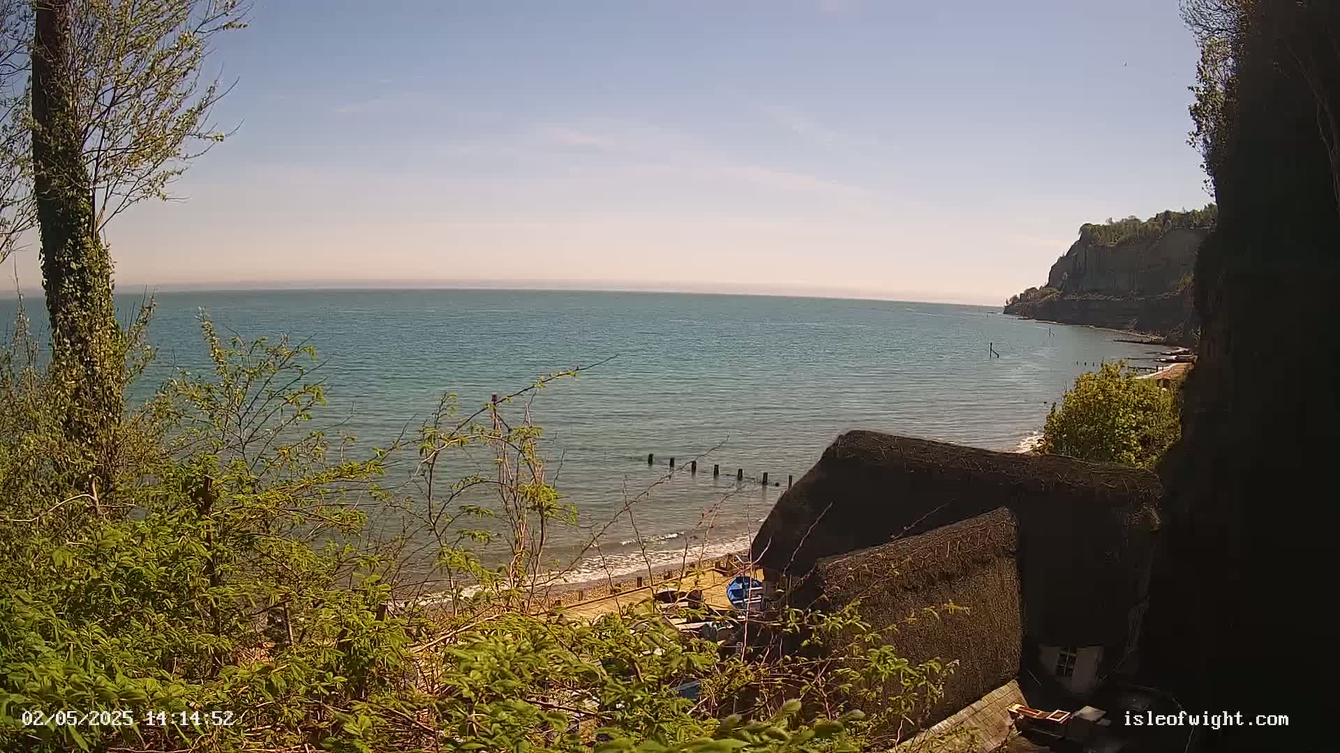 A tranquil seaside scene on a sunny day features a calm ocean, a sandy beach, a thatched roof structure, and verdant foliage overlooking the water and a distant cliff.