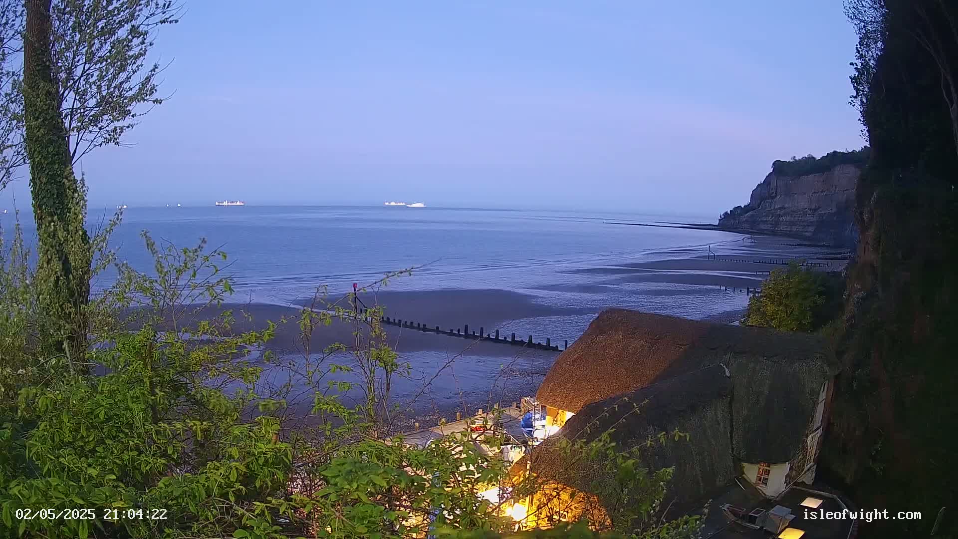 A tranquil seaside scene at dusk shows a calm sea with distant ships, a sandy beach, a low cliff, and a thatched-roof building partially obscured by vegetation.