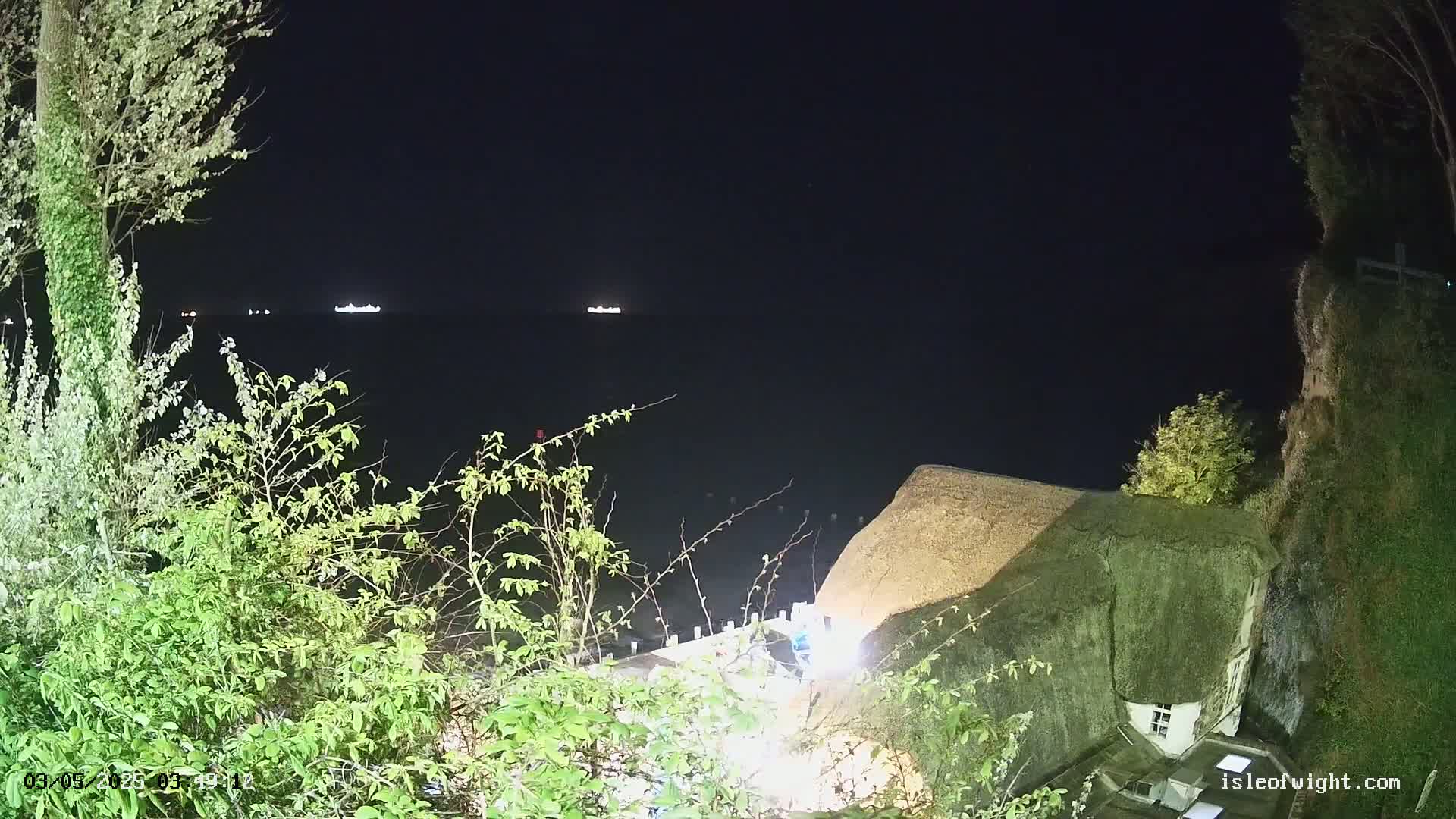 A nighttime view from a clifftop shows a dark body of water with distant lights, a thatched-roof house below, and vegetation in the foreground.