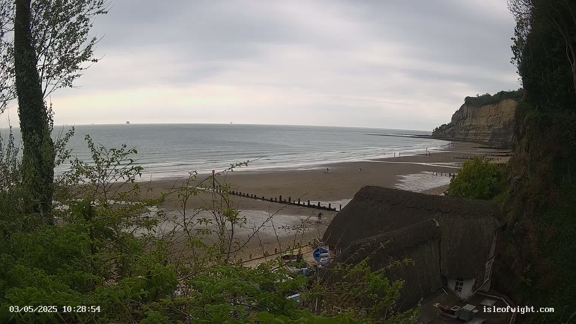 A sandy beach with a few people stretches out to a calm sea under a cloudy sky, partially obscured by vegetation and overlooking a thatched-roof building.