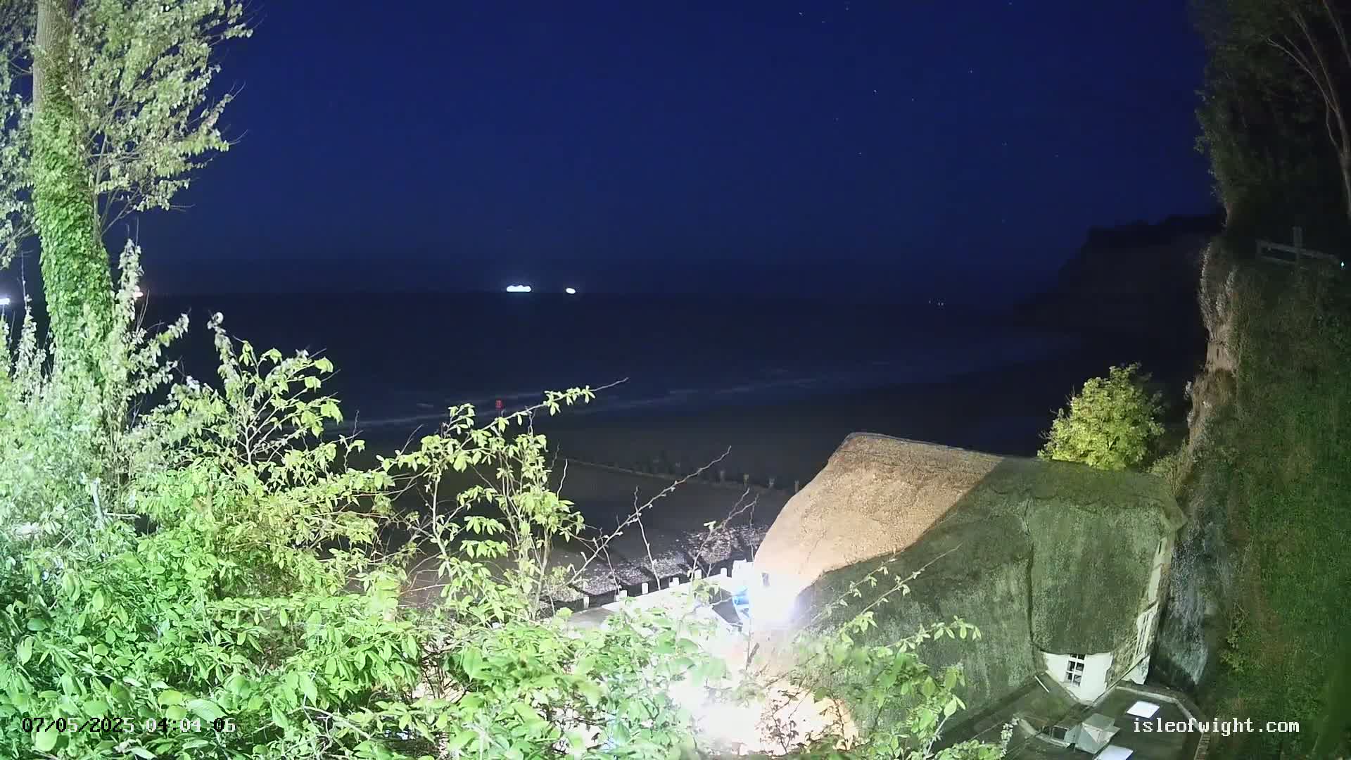 A thatched-roof building sits atop a cliff overlooking a dark, calm ocean under a night sky.