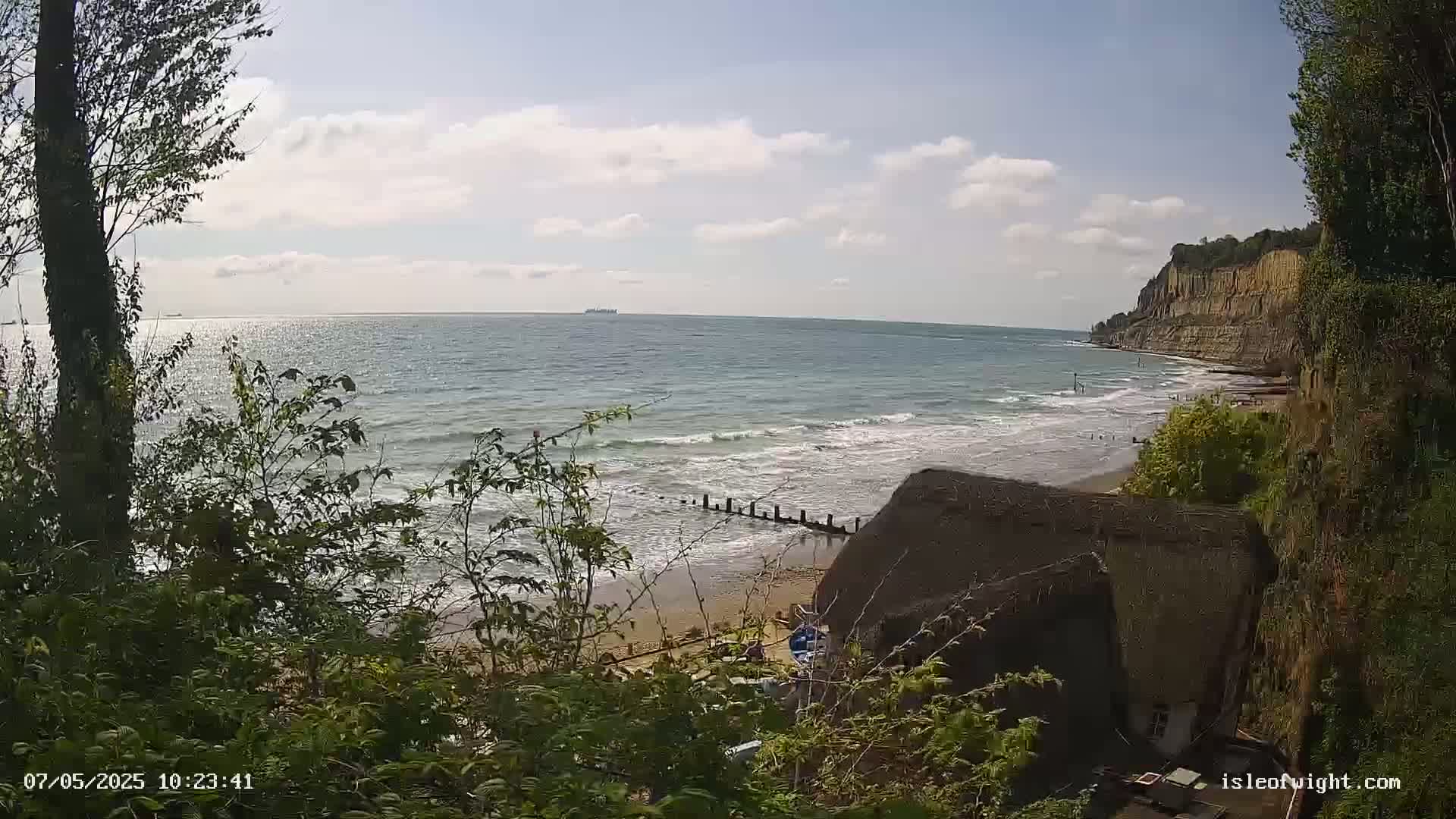 A sandy beach with gentle waves laps against a rocky cliff under a partly sunny sky; a thatched roof structure is partially visible in the foreground.