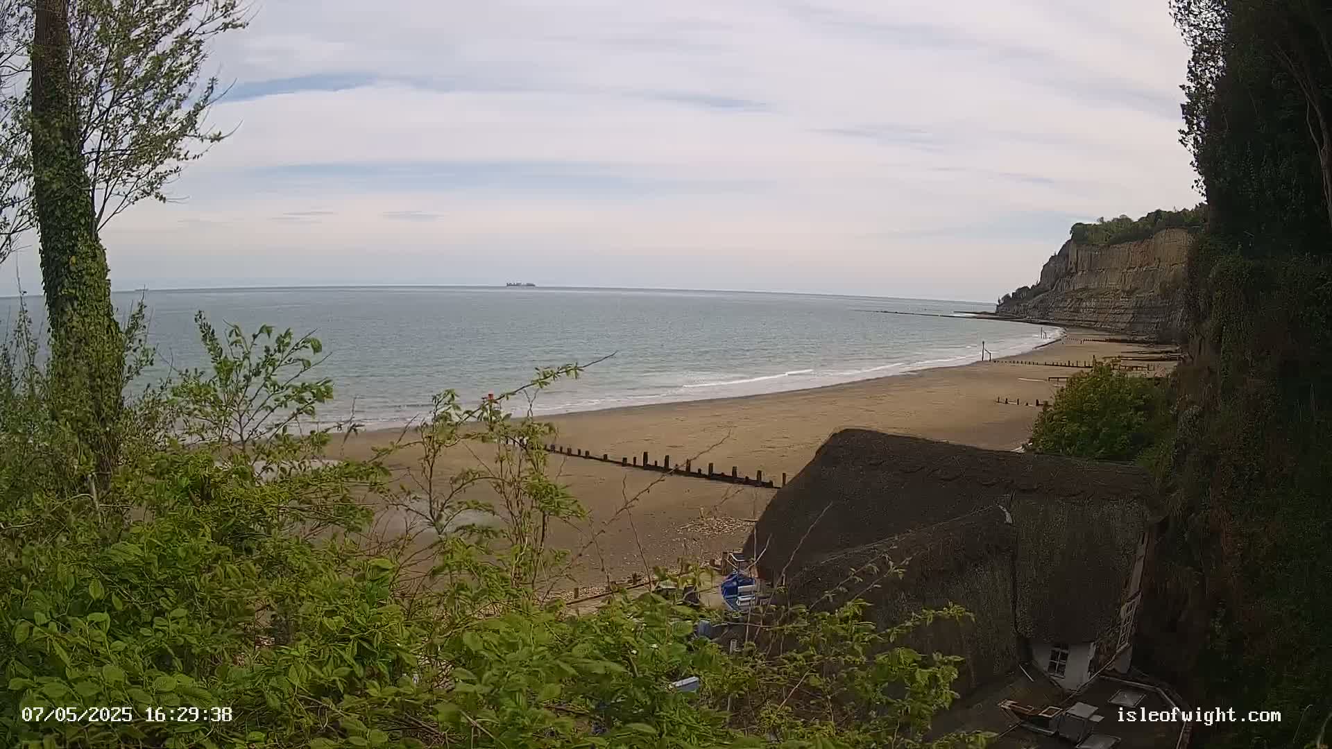 A sandy beach stretches below a rocky cliff, with a thatched-roof house nestled into the hillside, under a partly cloudy sky.