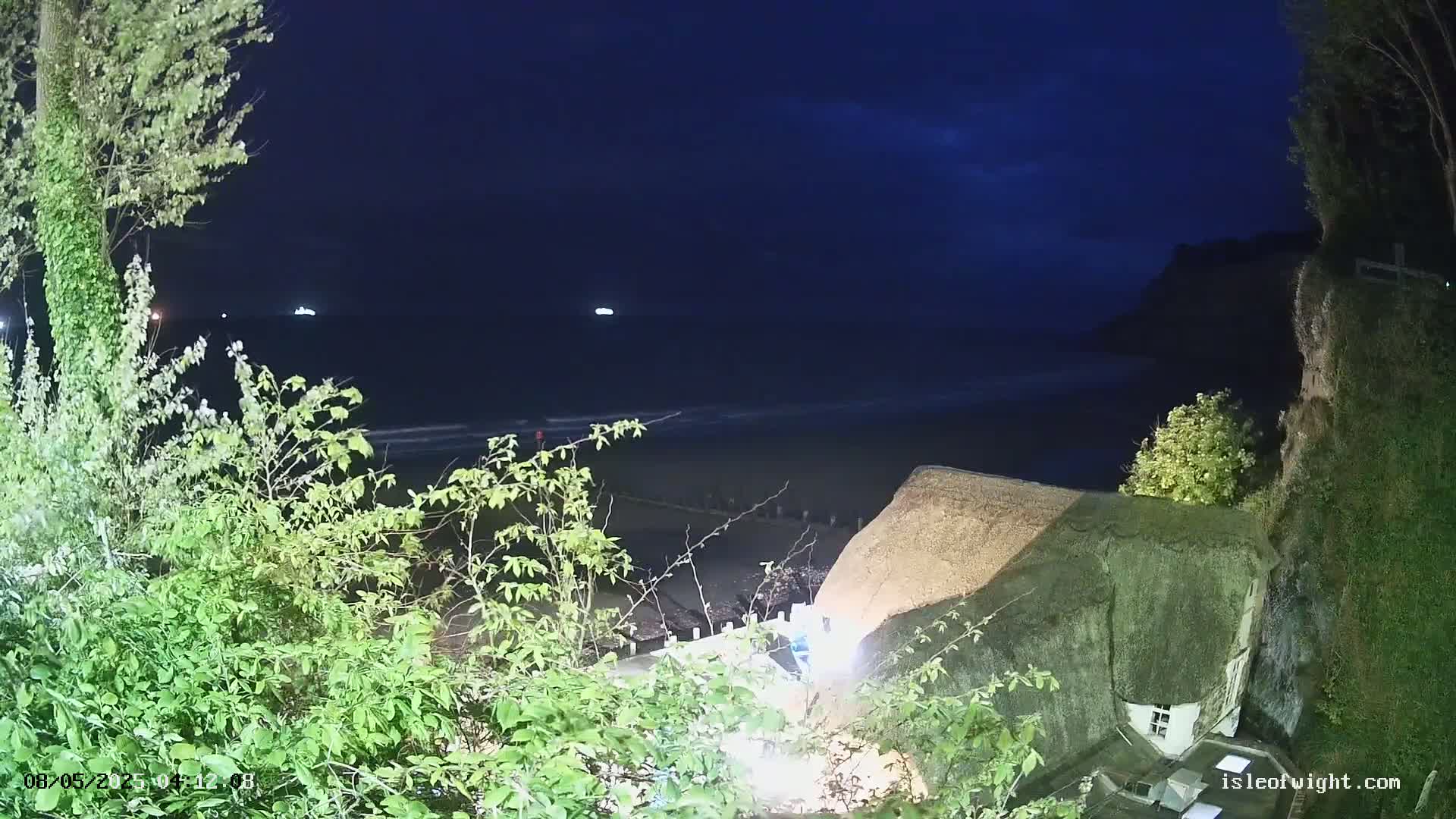 A thatched-roof house sits on a cliff overlooking a dark, nighttime beach and ocean with two distant lights visible on the water.