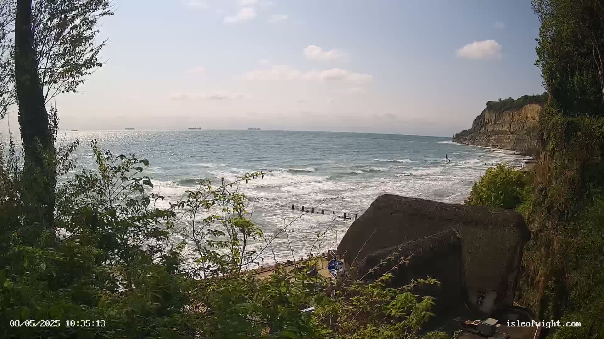 A partly sunny view of a rocky coastline with a partly cloudy sky, showing a choppy sea with waves breaking on the shore near a thatched roof structure partially obscured by vegetation.